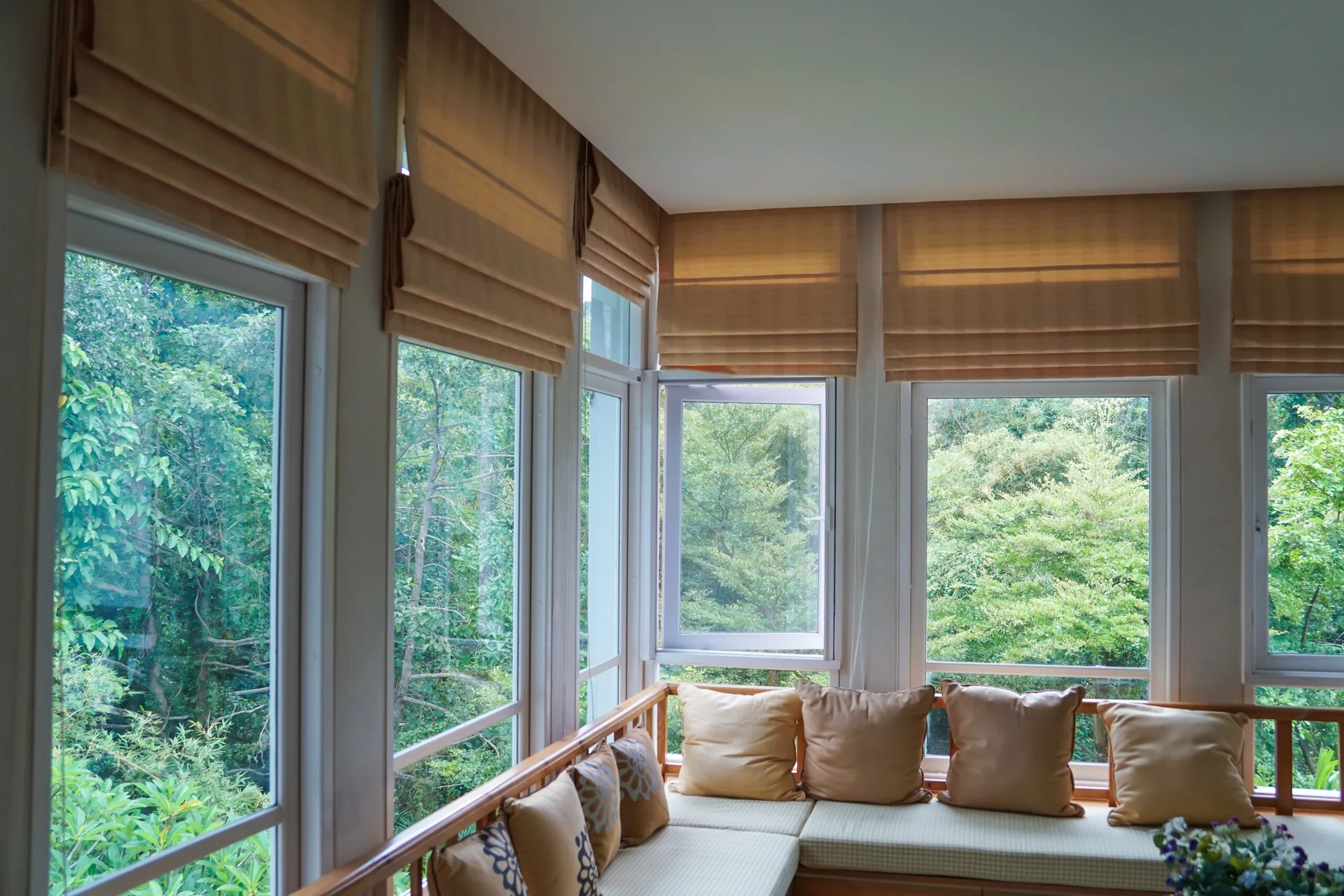 A built-in wooden corner bench with beige cushions and patterned pillows sits beneath large windows featuring matching fabric Roman blinds.
