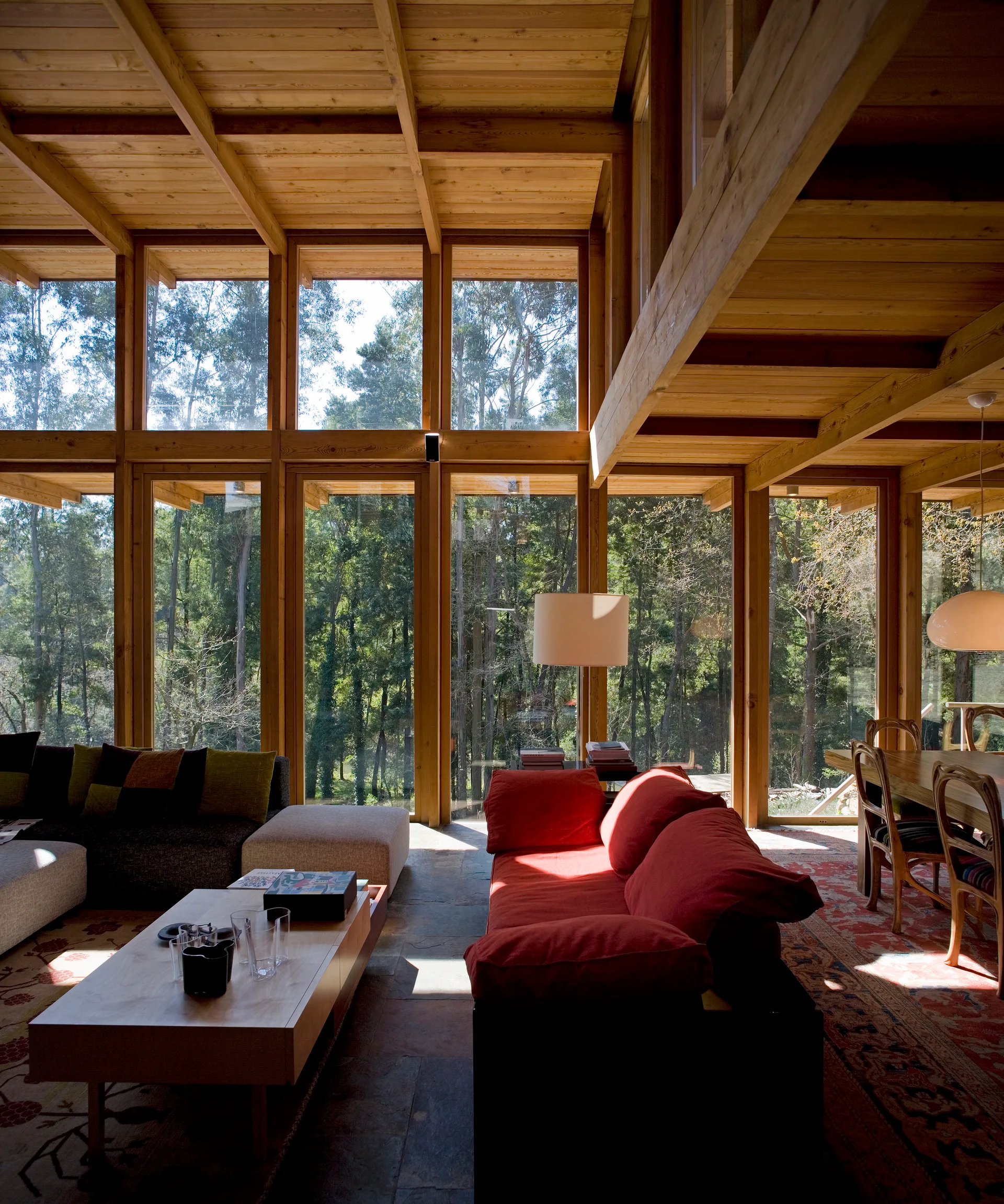 A rustic modern living room features floor-to-ceiling windows overlooking a forest, exposed wood beams, a red sofa, and stone flooring.