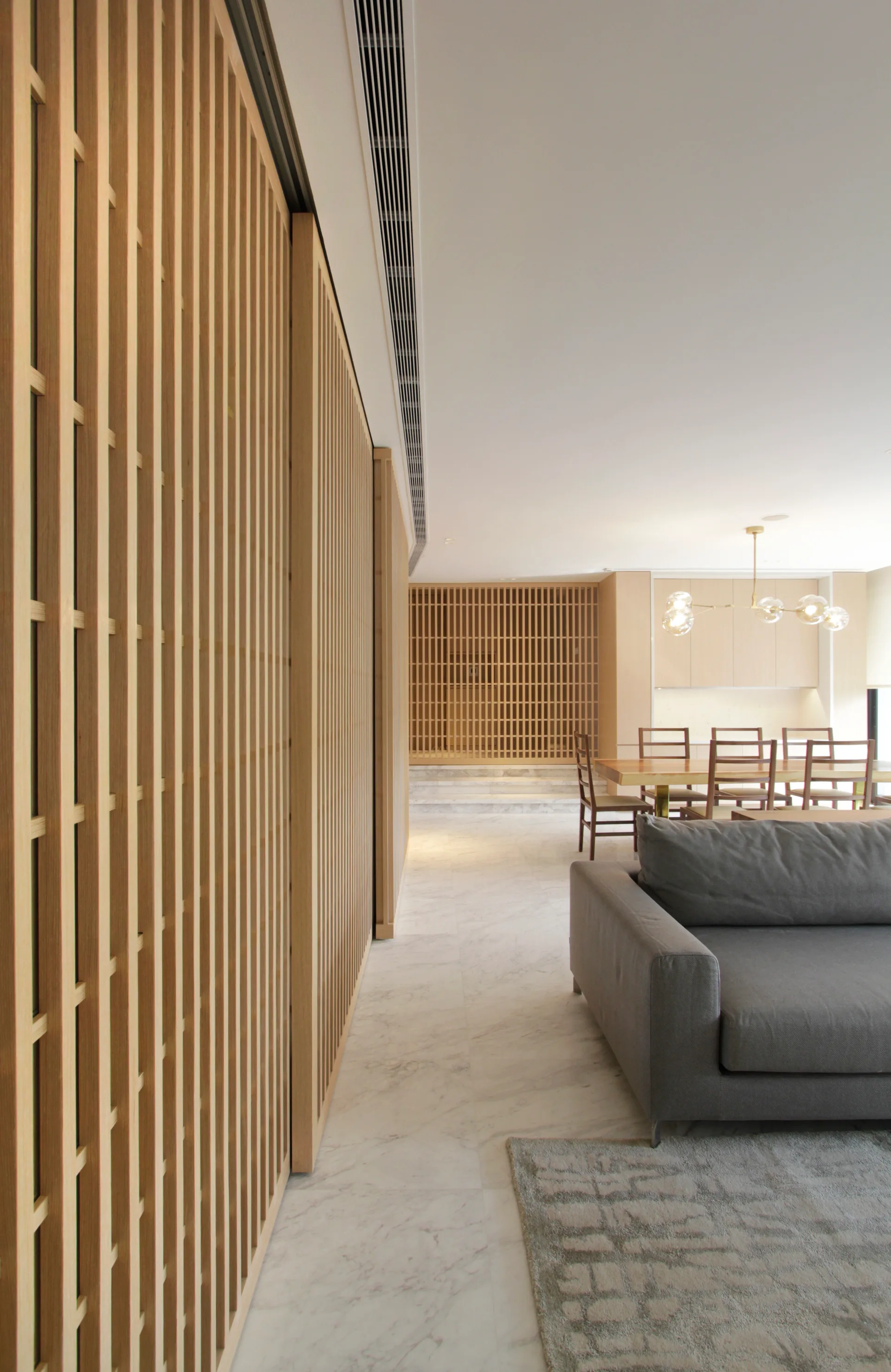 A modern open-plan living room features tall slatted wood partition walls, white marble floors, a gray sofa, and a distant dining area.