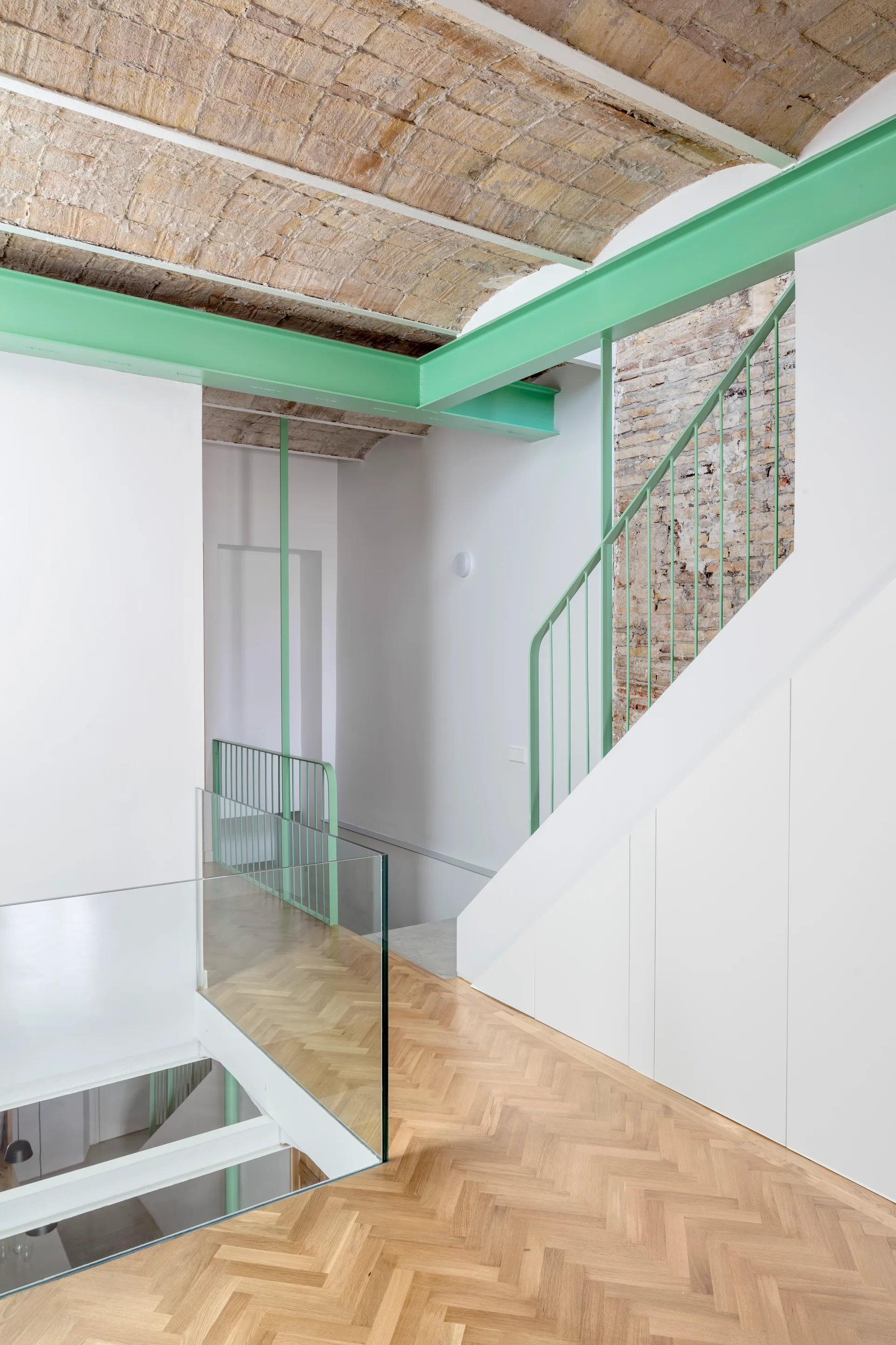 A modern staircase features herringbone wood floors, a vaulted brick ceiling, white under-stair storage, and bright mint green railings and support beams.