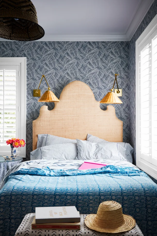 A bedroom featuring a tall arched rattan headboard against blue botanical wallpaper flanked by white plantation shutters and brass wall sconces.
