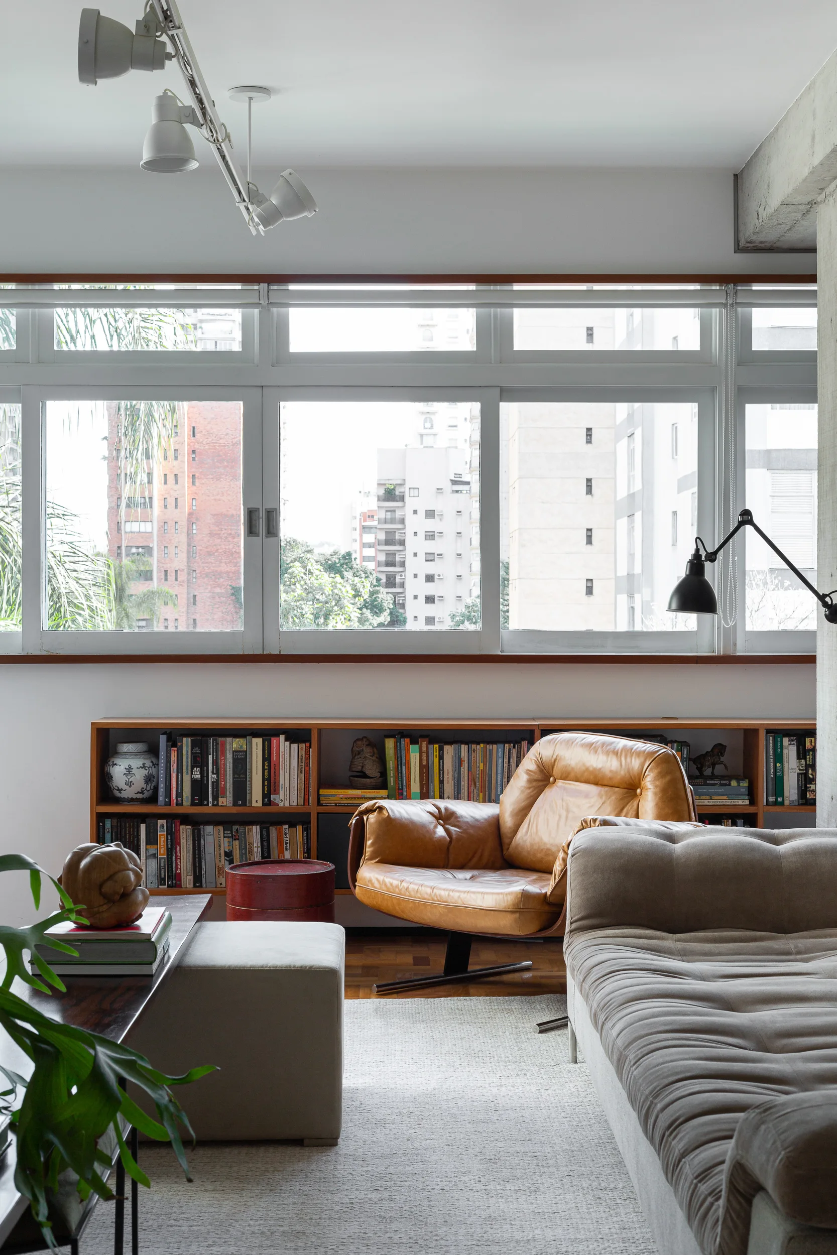 A modern living room features a tan leather lounge chair next to a low wood bookshelf situated beneath a large window.