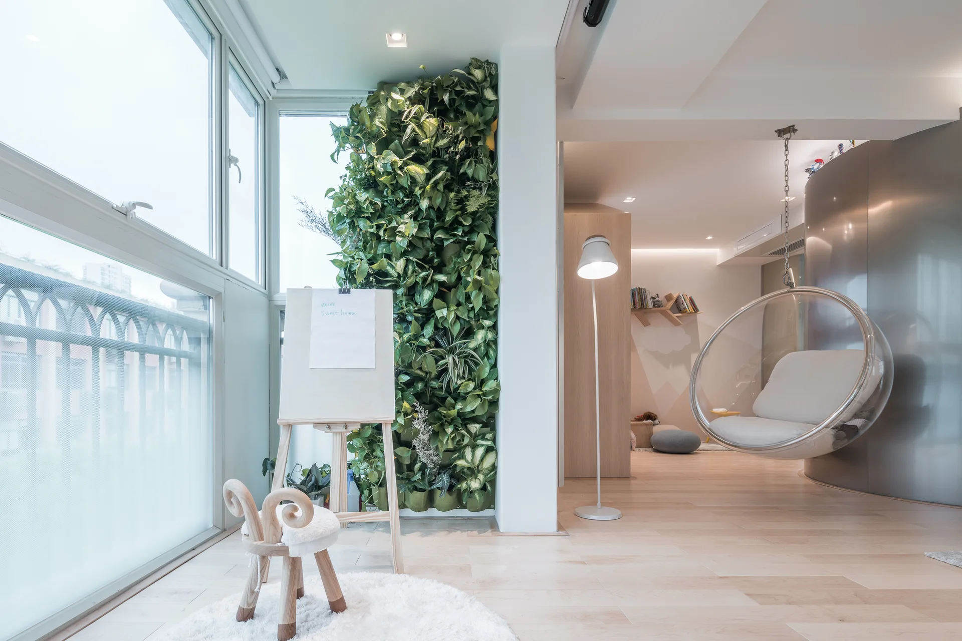 A bright modern playroom features a vertical garden wall, a transparent hanging bubble chair, and a child-sized wooden easel on light floors.
