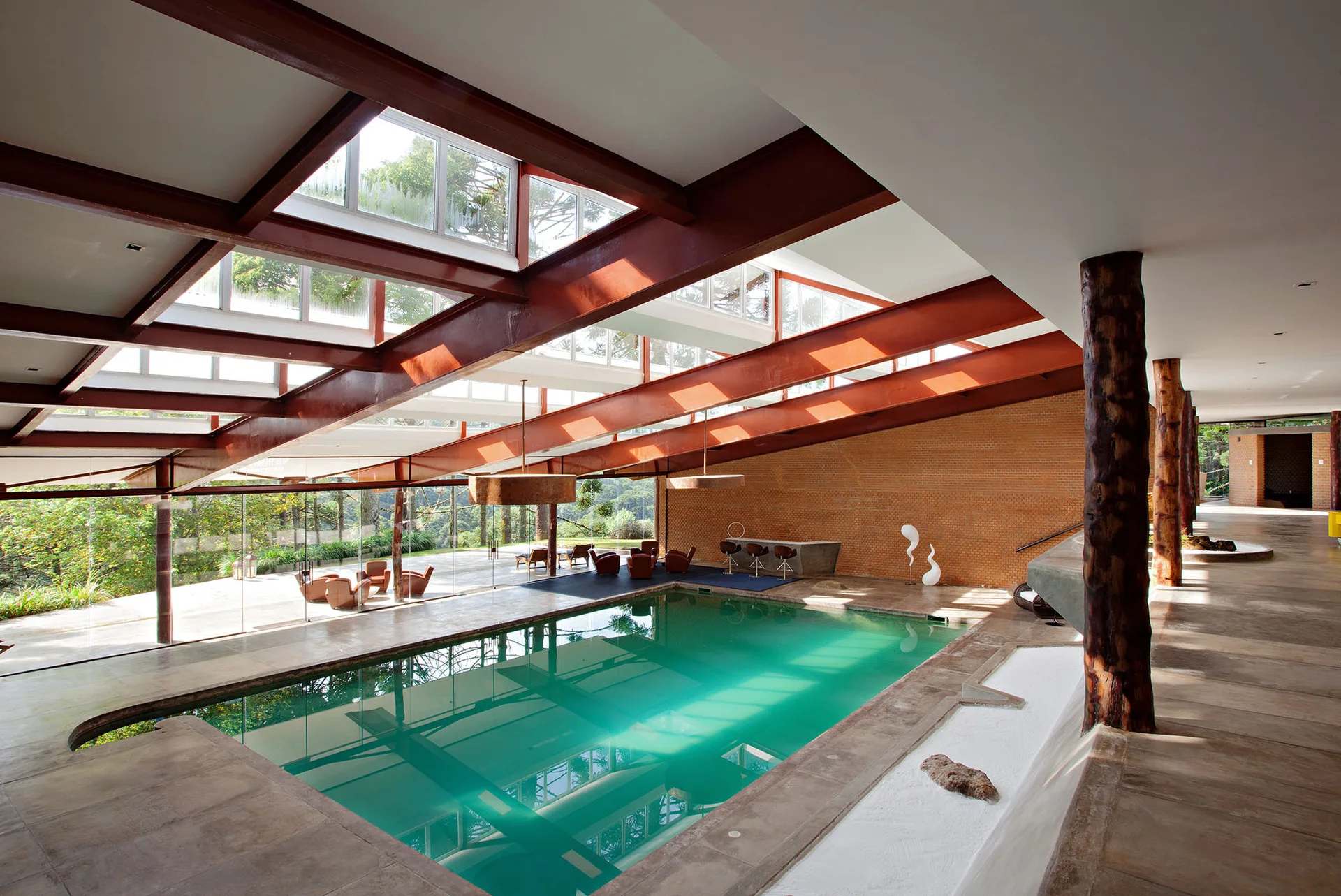 A spacious indoor swimming pool featuring exposed steel ceiling beams, large skylights, floor-to-ceiling glass walls, and rustic log columns.