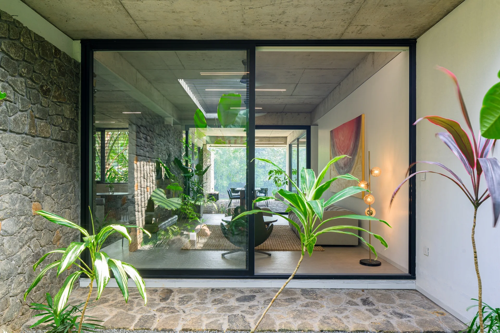 A modern indoor-outdoor living room seen through a sliding glass door, featuring a stone wall, a concrete ceiling, a sofa, and green plants.