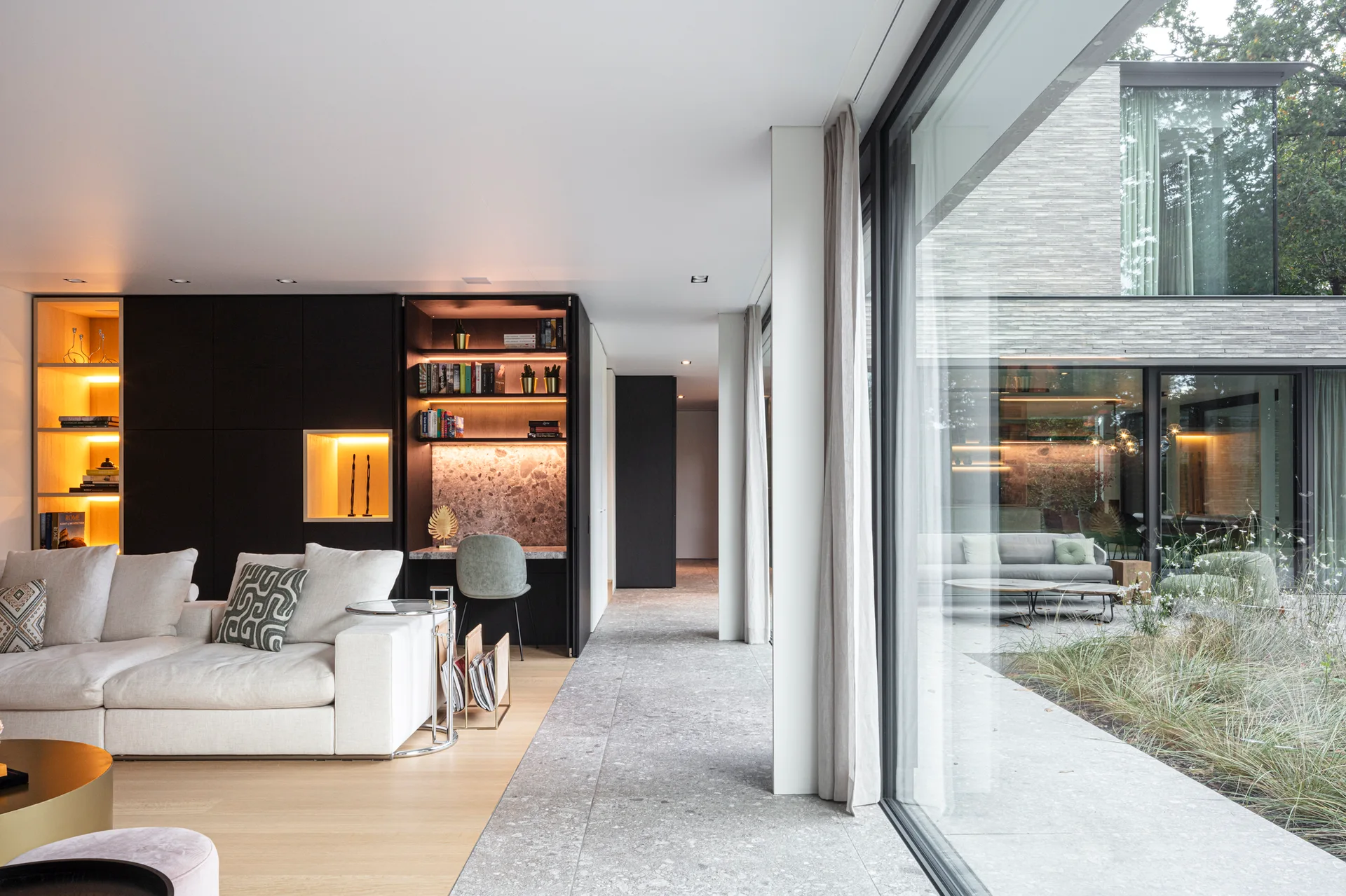 A modern living room features a cream sectional sofa, integrated black cabinetry with a lit desk nook, and large glass windows facing a courtyard.