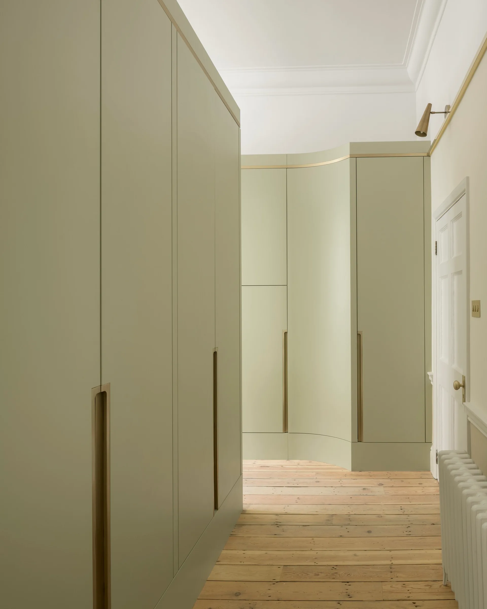 A hallway features custom sage green wardrobes with recessed brass pulls and light wood plank floors.