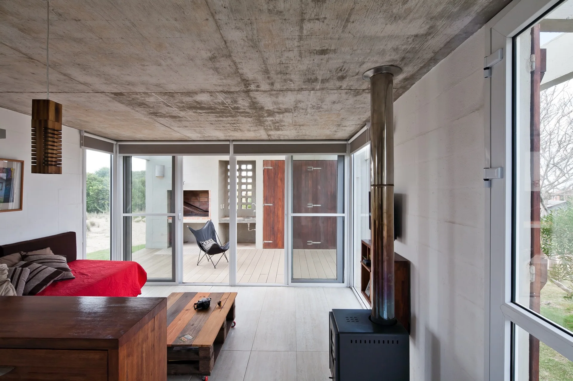 An industrial living room featuring a raw concrete ceiling, a black wood stove with a metal flue, and glass sliding doors that open to a wooden deck.