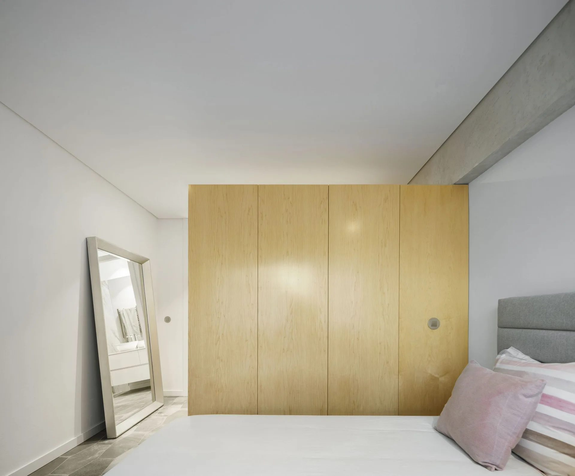 A minimalist bedroom features a large light wood wardrobe, a bed with soft pink pillows, and a large floor mirror leaning against a white wall.