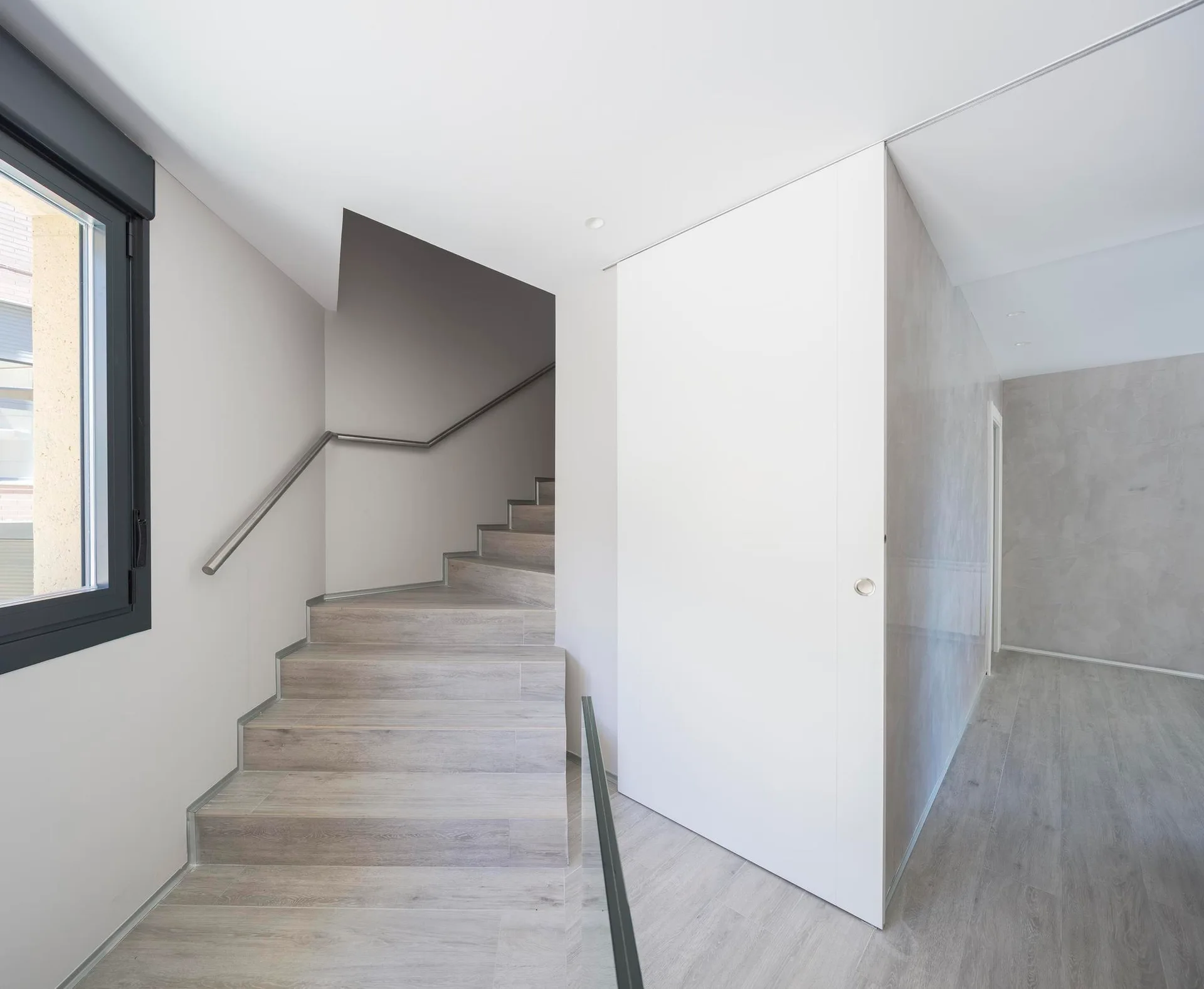 A minimalist interior staircase with light wood steps, a metallic handrail, and a large white sliding wall panel.