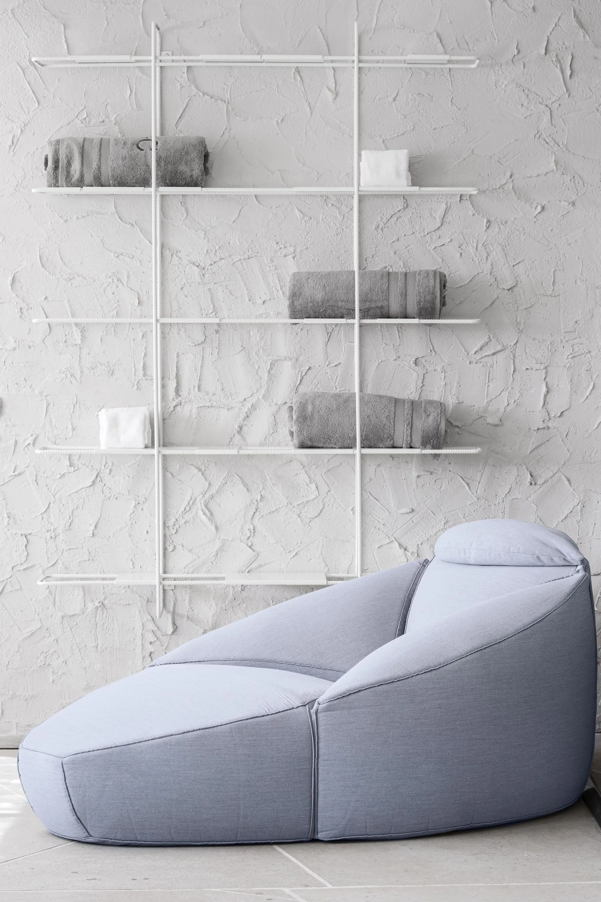 A low-profile light blue lounge chair sits on a tile floor in front of a heavily textured white wall with a white wire shelving unit.