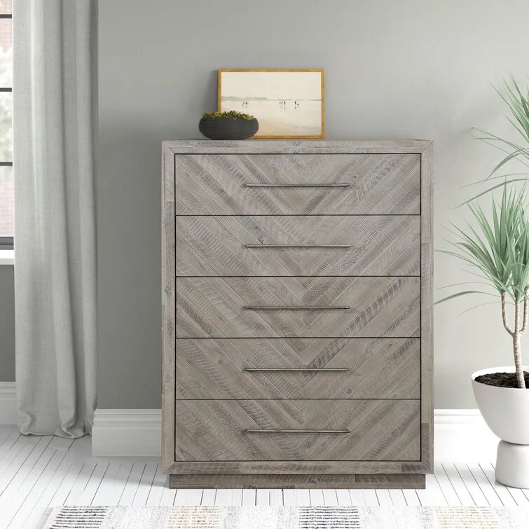 A five-drawer grey wooden dresser with a herringbone pattern on the drawer fronts stands next to a white planter.