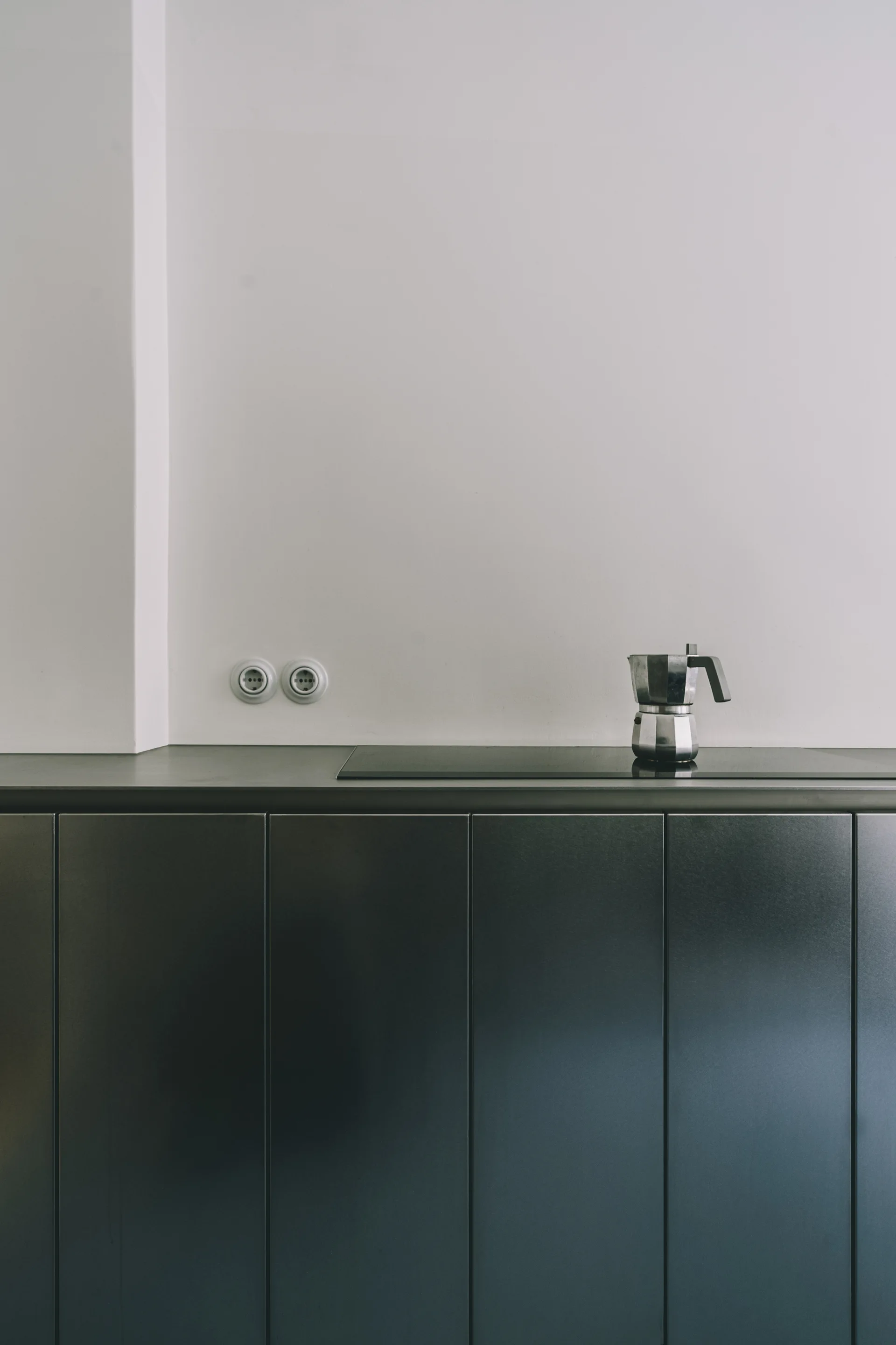 Minimalist kitchen counter with matte metallic grey cabinets, a black stovetop holding a moka pot, and two circular wall outlets.