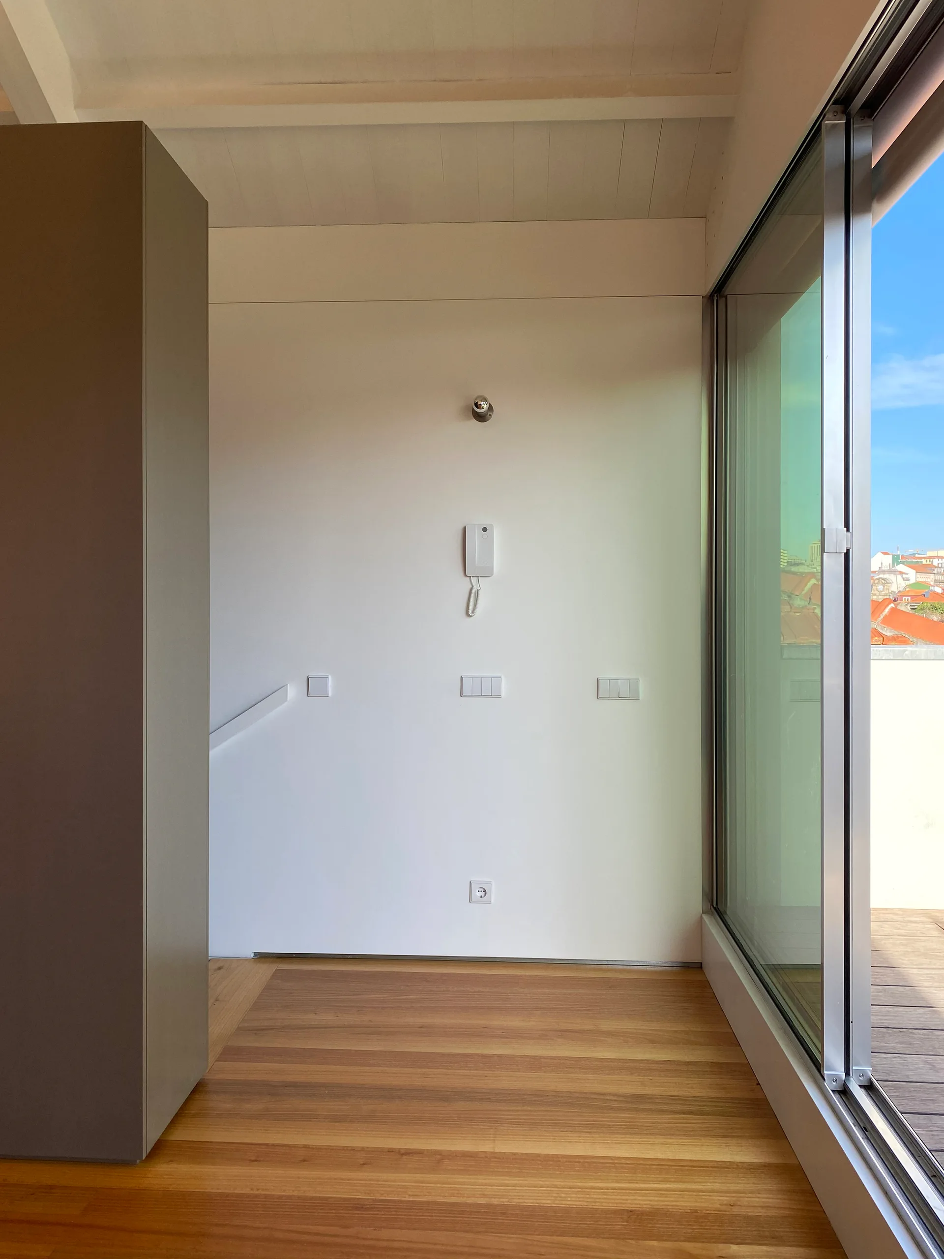 A minimalist white stair landing features a sliding glass door opening to a wooden terrace, alongside a wall with an intercom and light switches.