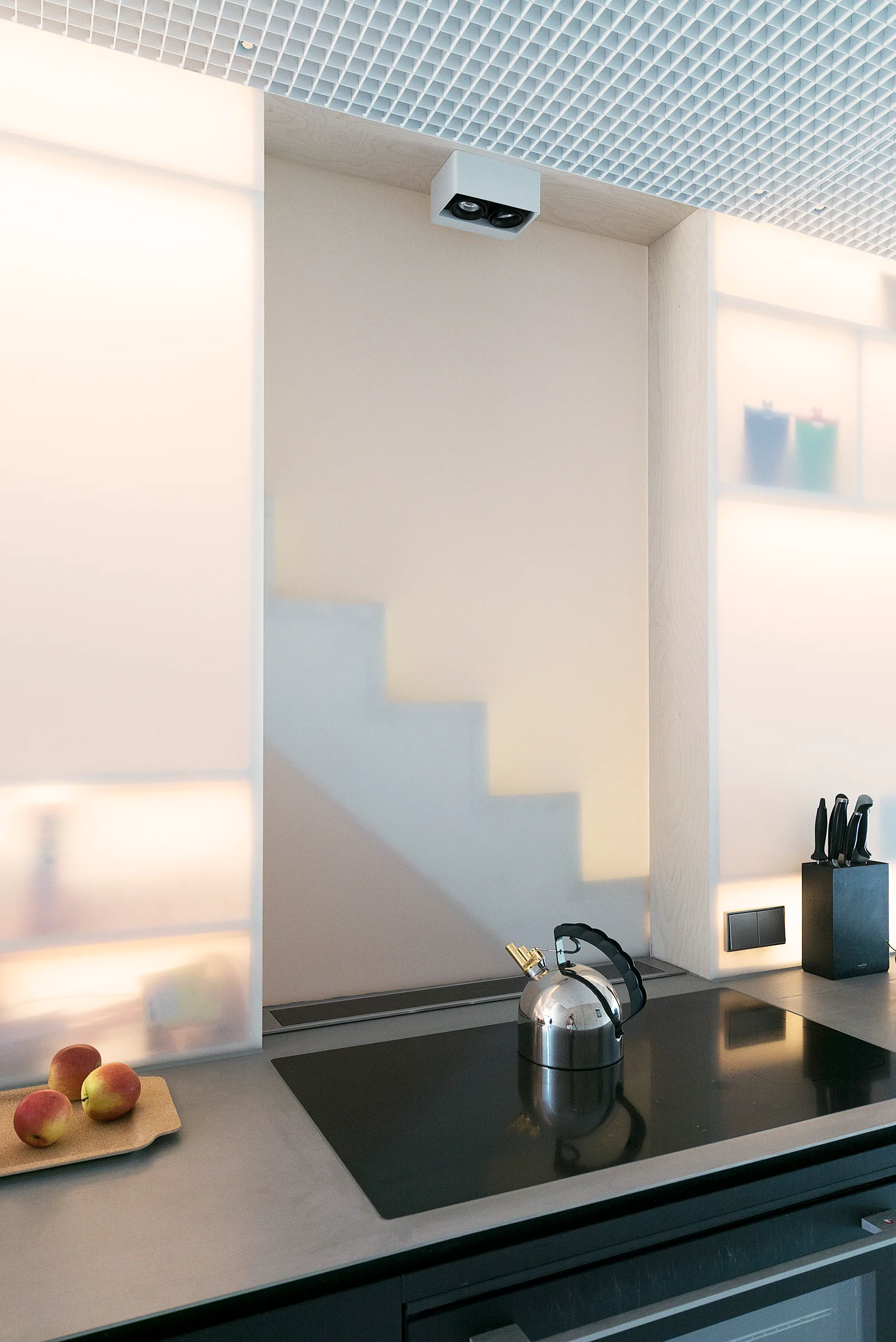 A modern kitchen counter features an induction cooktop with a metal kettle, surrounded by glowing backlit frosted glass panels and a white grid ceiling.