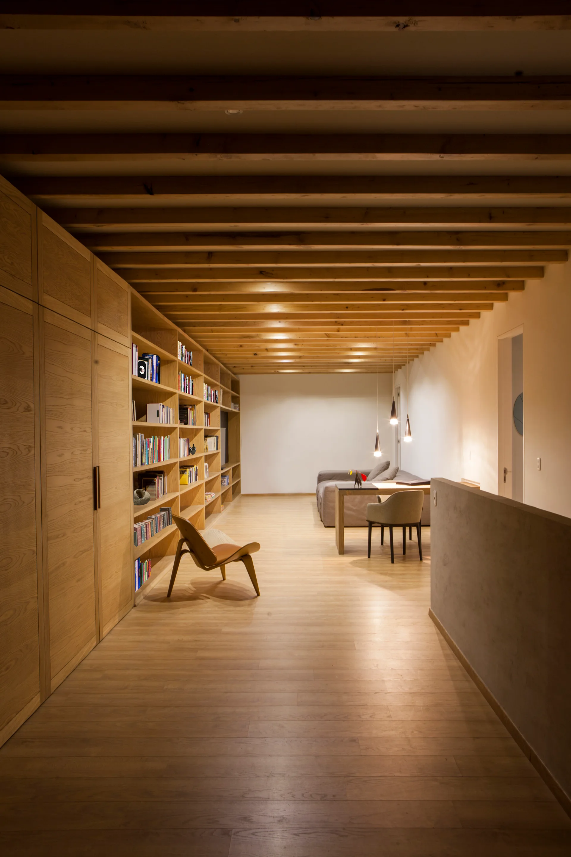A minimalist room features a large built in wooden bookshelf, exposed timber ceiling beams, matching wood floors, and a distant seating area.