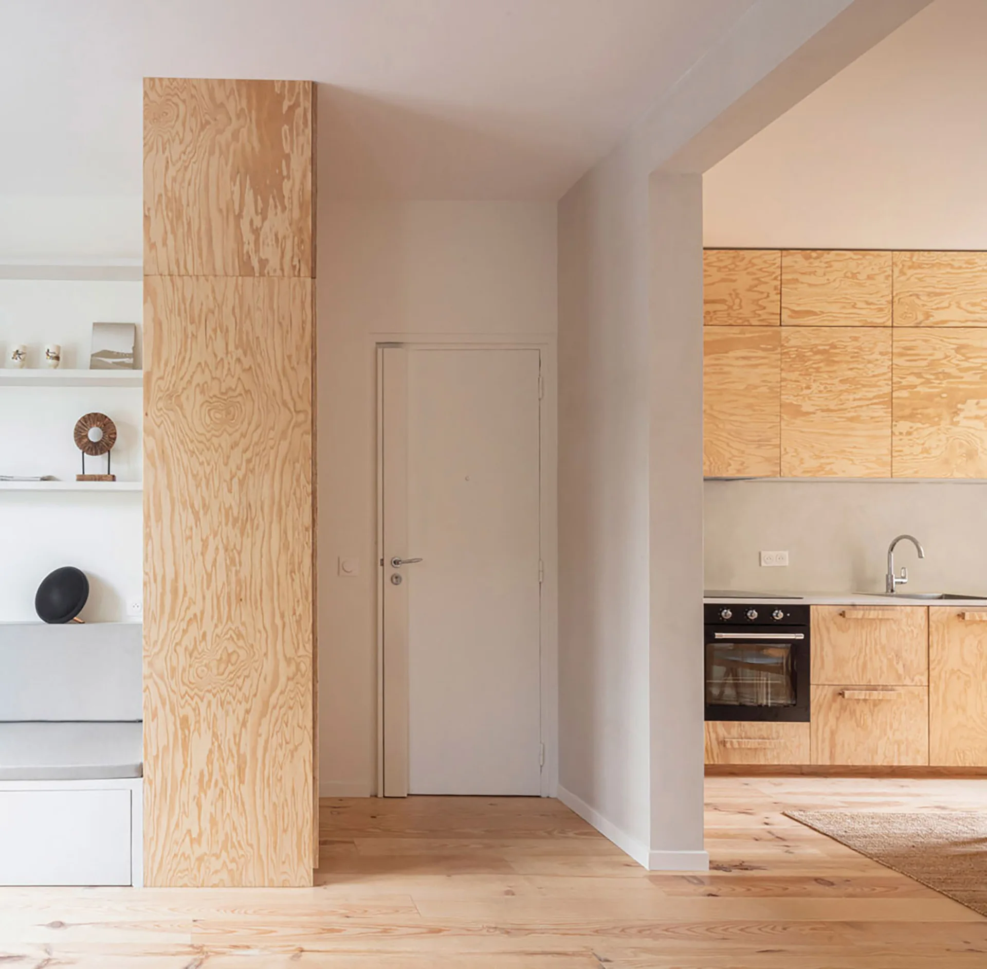 A bright interior features a modern kitchen with natural plywood cabinets, an adjacent white door, and a matching plywood-clad partition column.