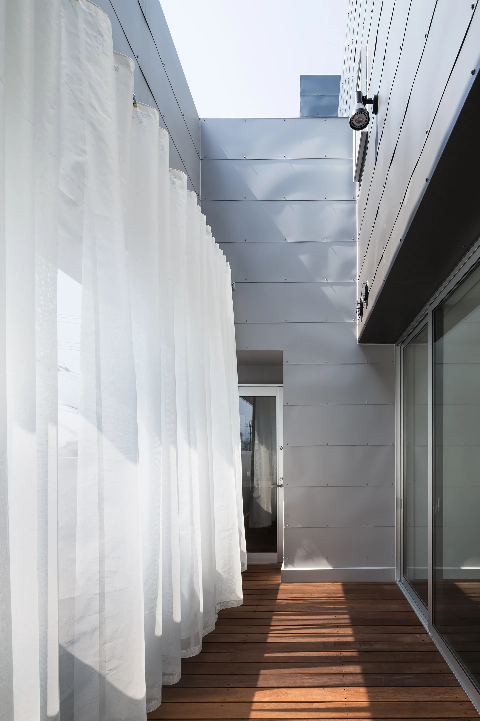 A narrow modern balcony with wood plank flooring, a glass sliding door, grey metal wall siding, and long sheer white curtains.