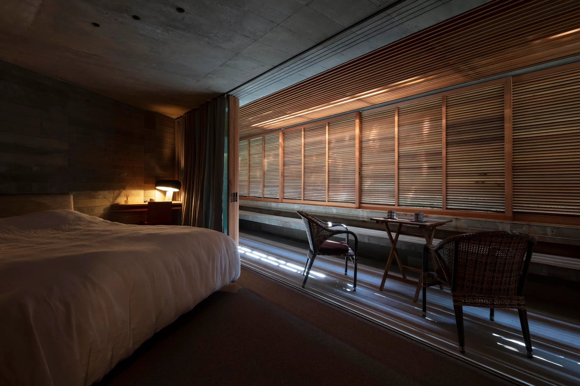 A bedroom features an exposed concrete ceiling, a low bed, and warm wooden louvered shutters behind two rattan armchairs.