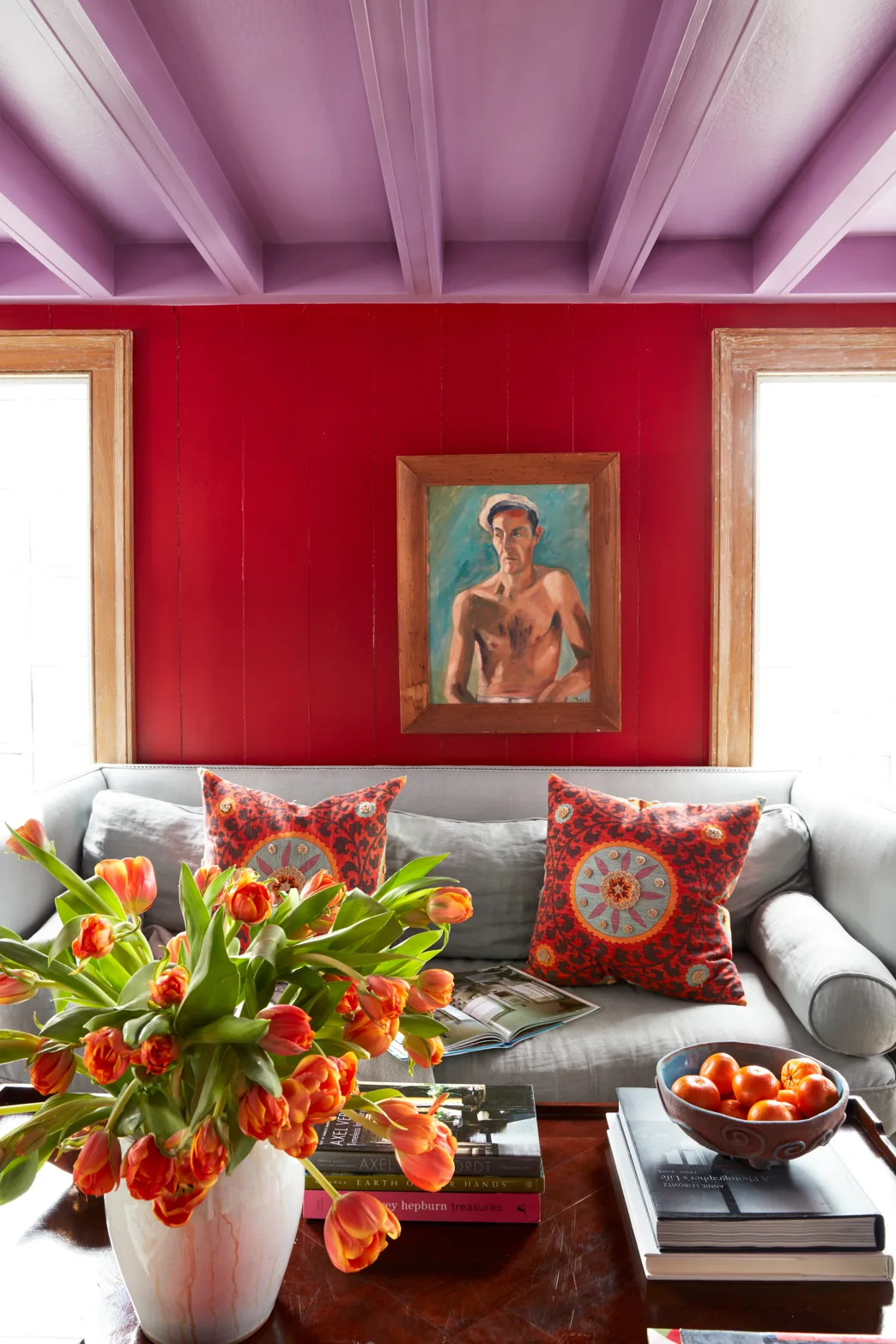 A gray sofa sits against a red wood-paneled wall beneath a light purple beamed ceiling in an eclectic living room.