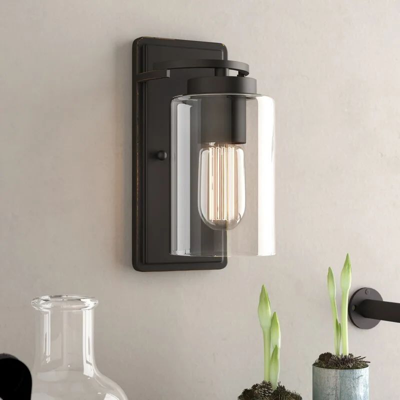 A matte black industrial wall sconce with a cylindrical clear glass shade and an illuminated warm bulb is mounted on a light beige wall.