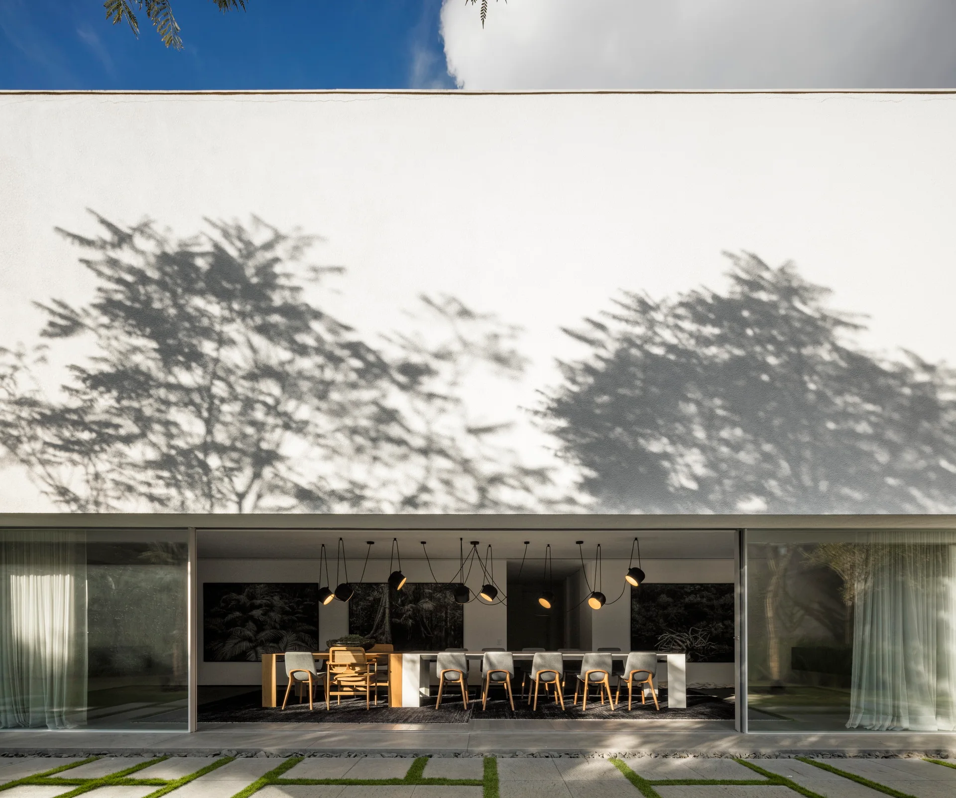 A modern indoor-outdoor dining room with a long table and black pendant lights sits beneath a large white wall covered in tree shadows.