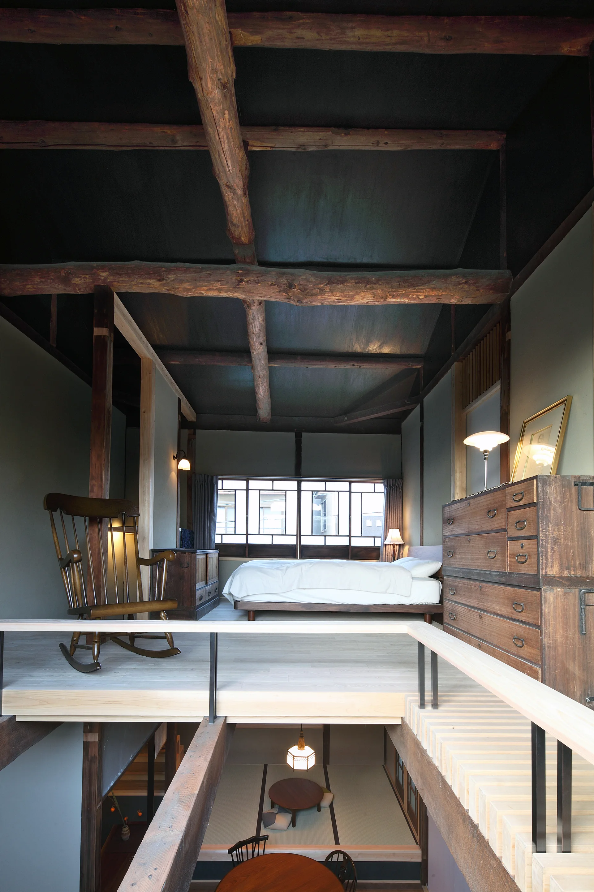 A split-level loft bedroom features a platform bed, a vintage wooden dresser, and a rocking chair beneath heavy exposed ceiling log beams.