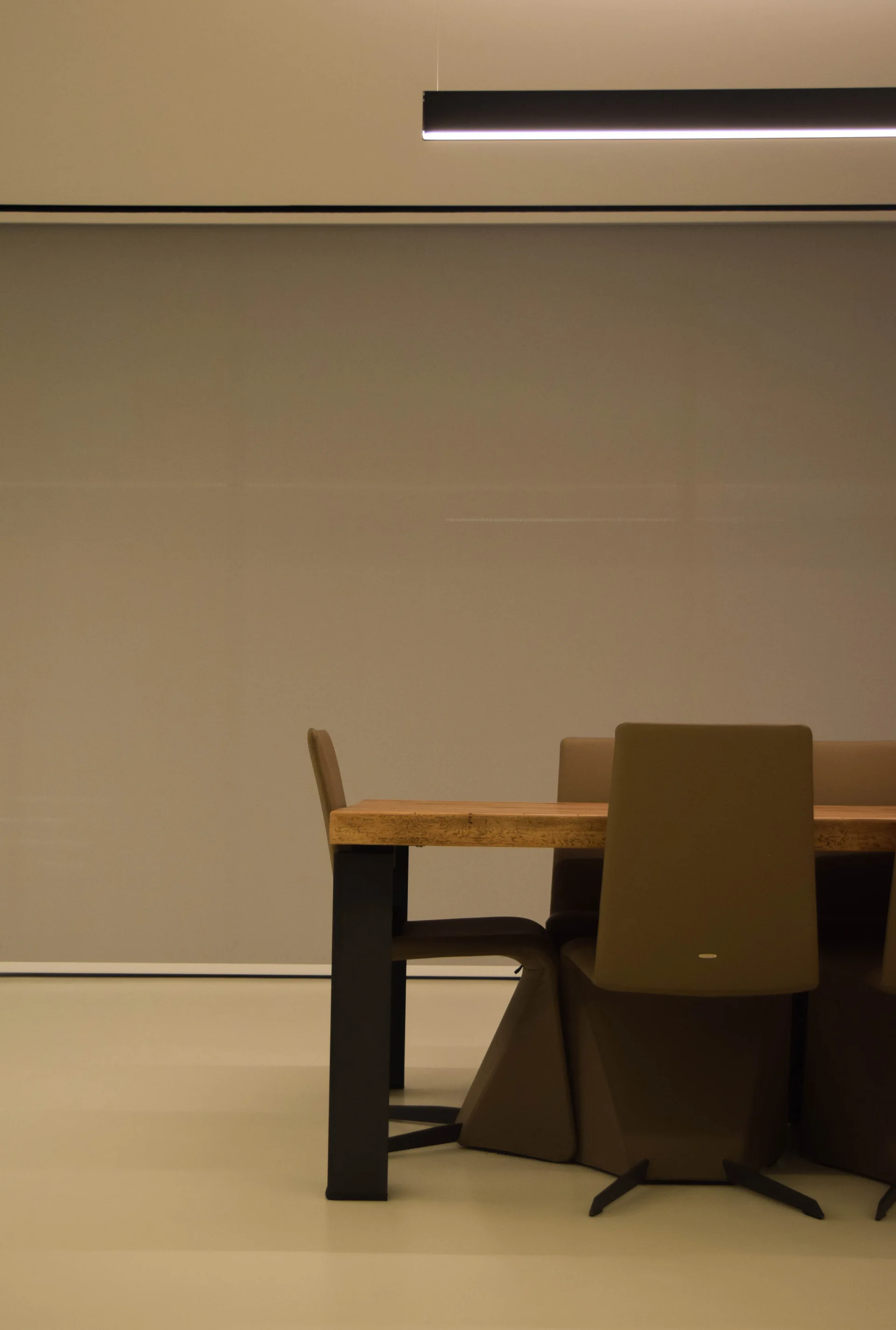 A minimalist dining room features a natural wood table with taupe upholstered chairs beneath a sleek black linear pendant light against a grey wall.
