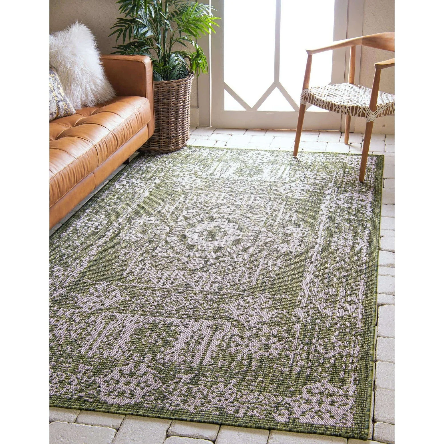 A green and cream medallion pattern area rug rests on a stone paver floor in a sunlit room alongside a tan leather sofa and a wooden accent chair.
