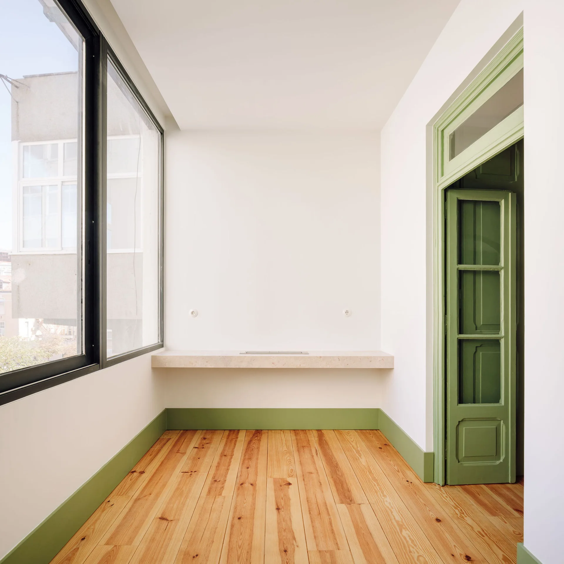 A minimalist white room featuring natural light from large windows, light wood floors, sage green trim, and a floating stone shelf.