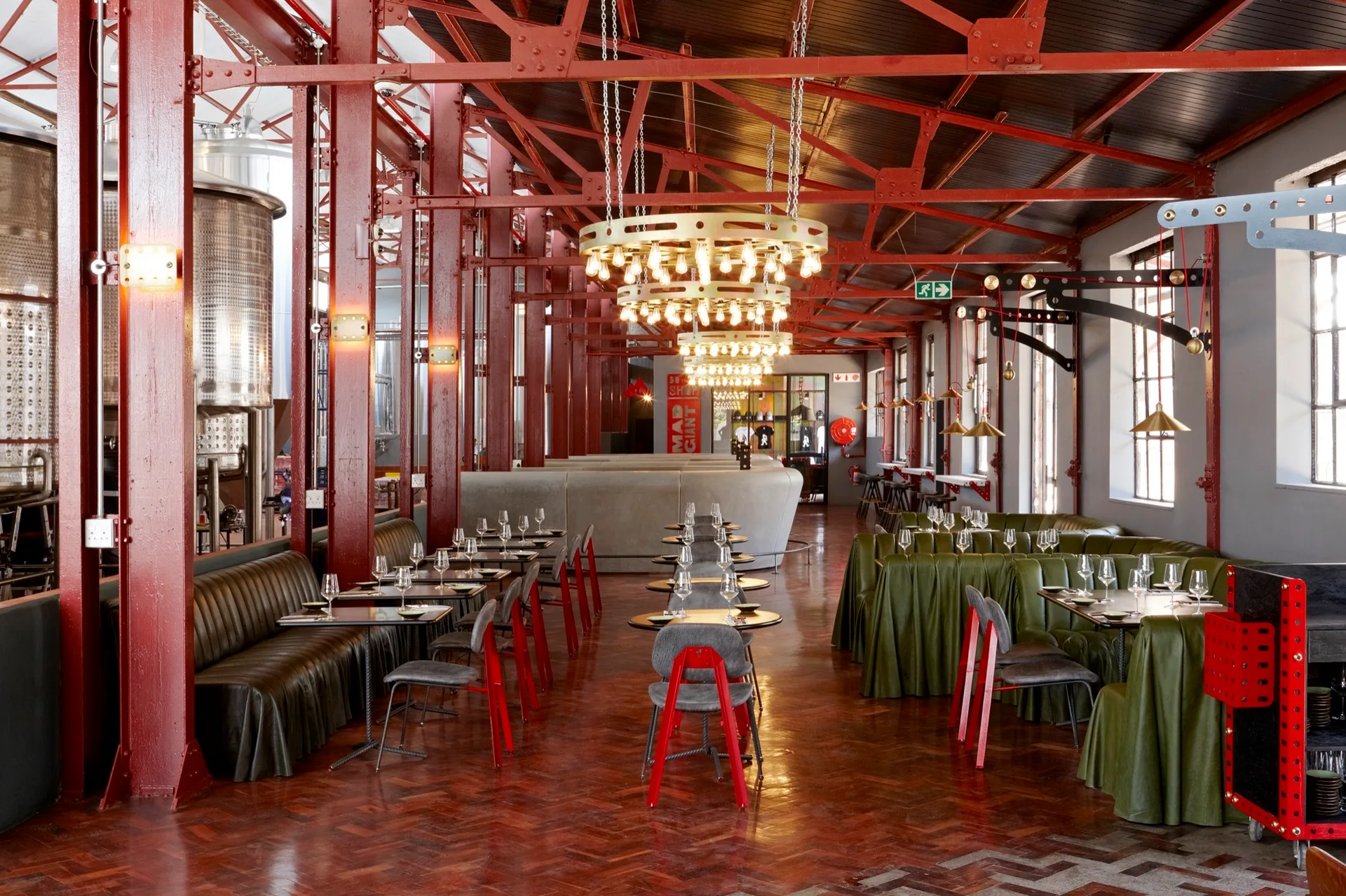 An industrial restaurant dining room features prominent red steel beams, olive green banquette seating, and large circular gold chandeliers over wooden tables.