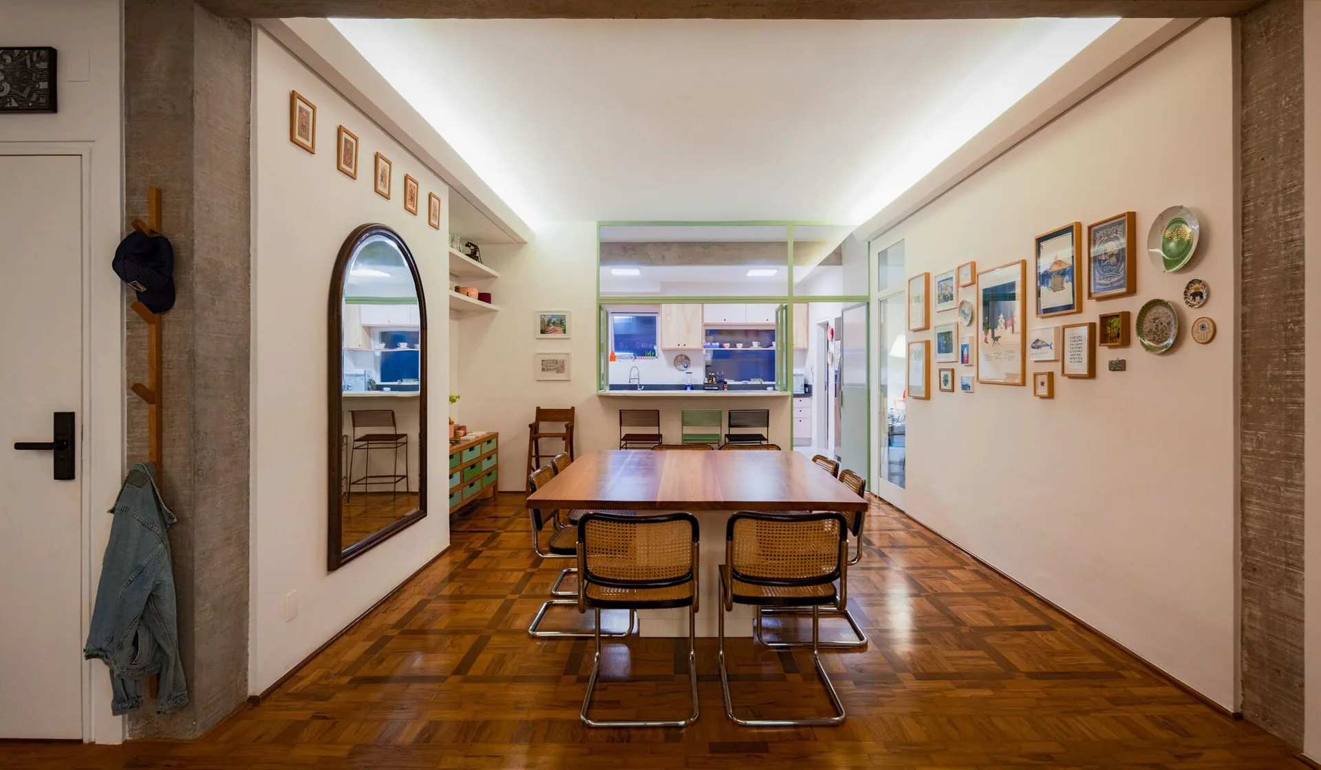 A dining room features a wood table with cane chairs, parquet flooring, and a green-framed window looking into the kitchen.
