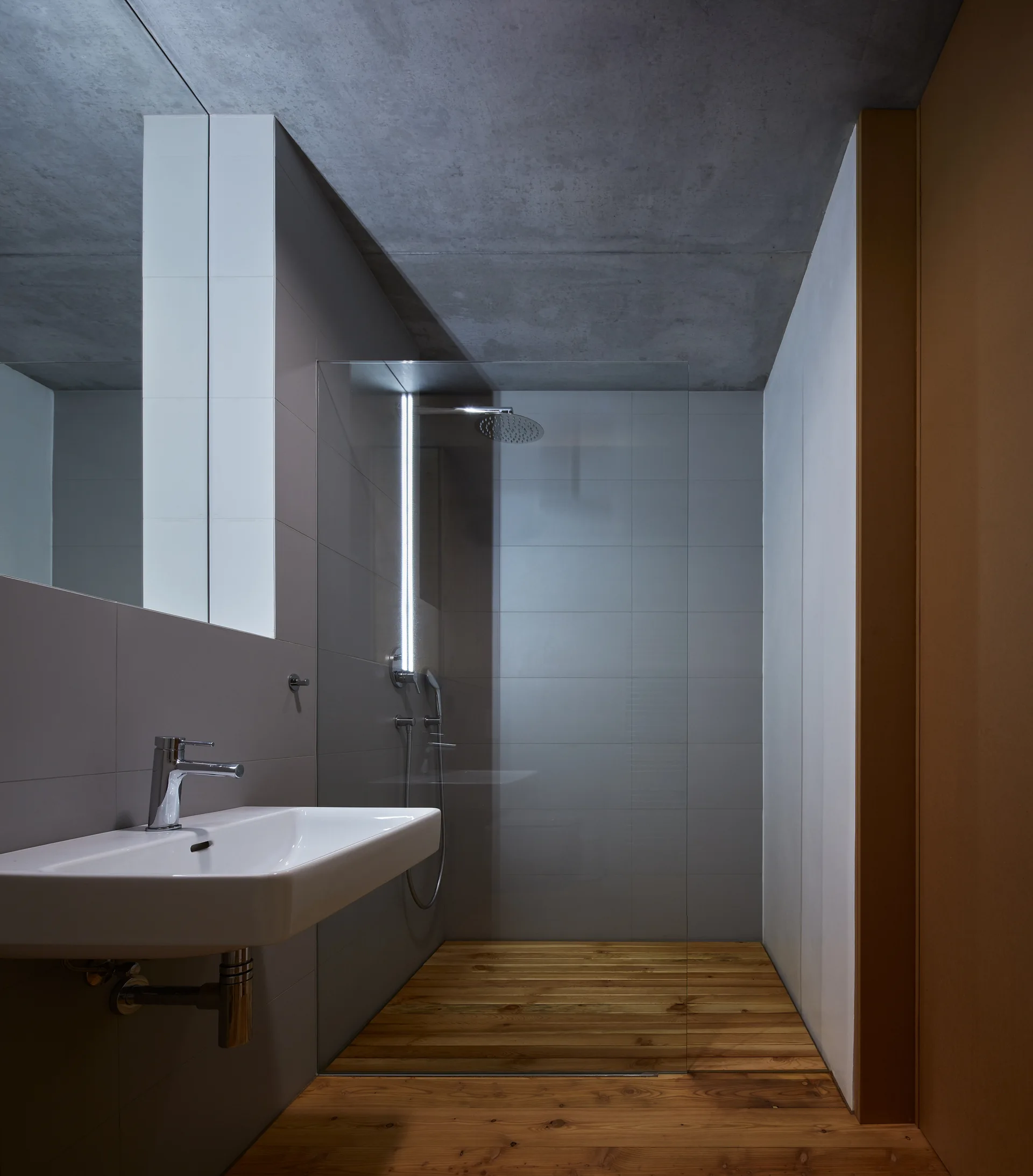 A minimalist bathroom features a wall-mounted sink, raw concrete ceiling, and a glass shower enclosure with warm wood plank flooring.