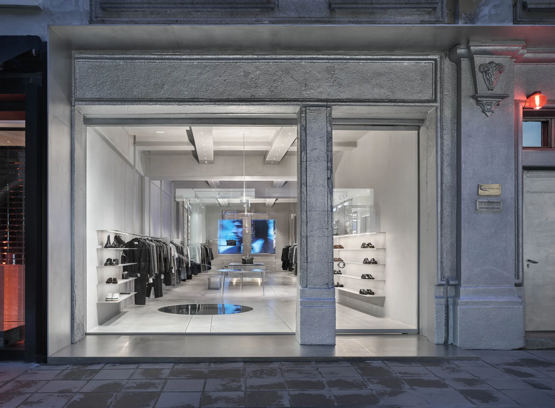 A minimalist retail store interior with concrete-like walls and under-shelf lighting is viewed through a large glass storefront window.
