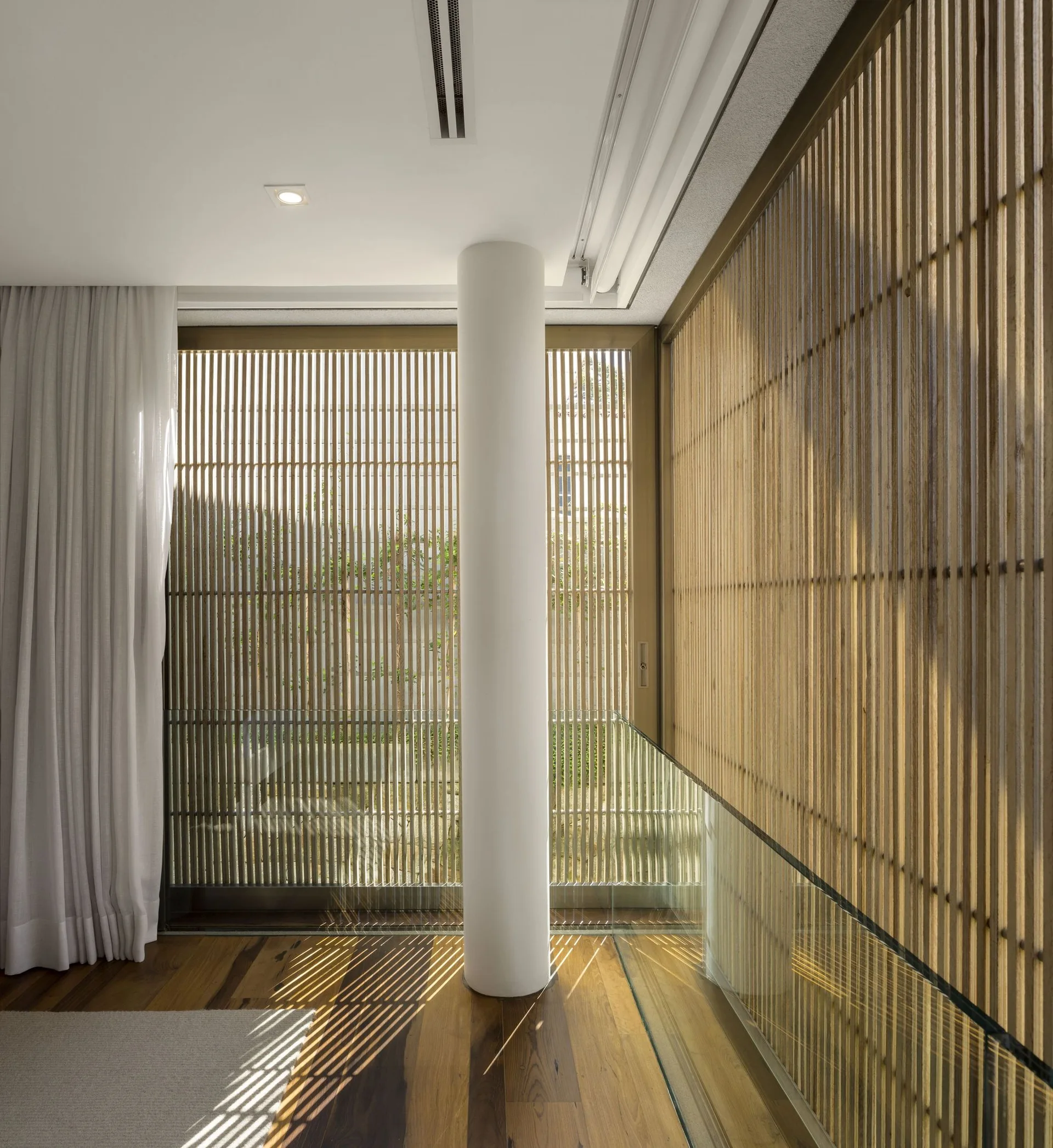A modern room features a floor-to-ceiling vertical wood slat wall, polished hardwood flooring, a white structural column, and a glass railing.