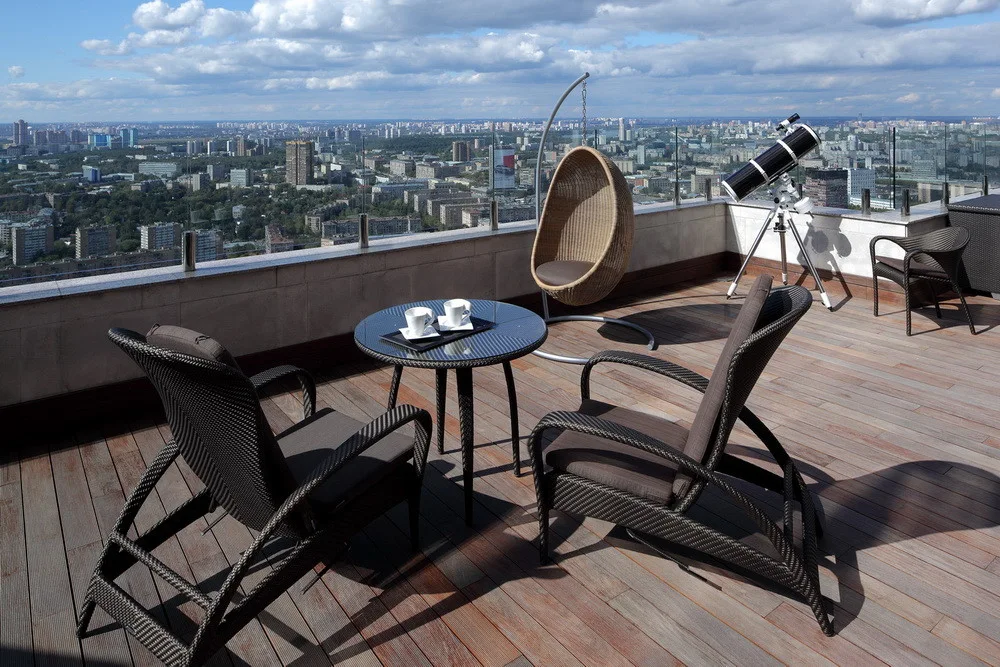 Dark wicker armchairs and a small glass bistro table sit on a wooden rooftop terrace with a hanging egg chair and a telescope overlooking a city skyline.