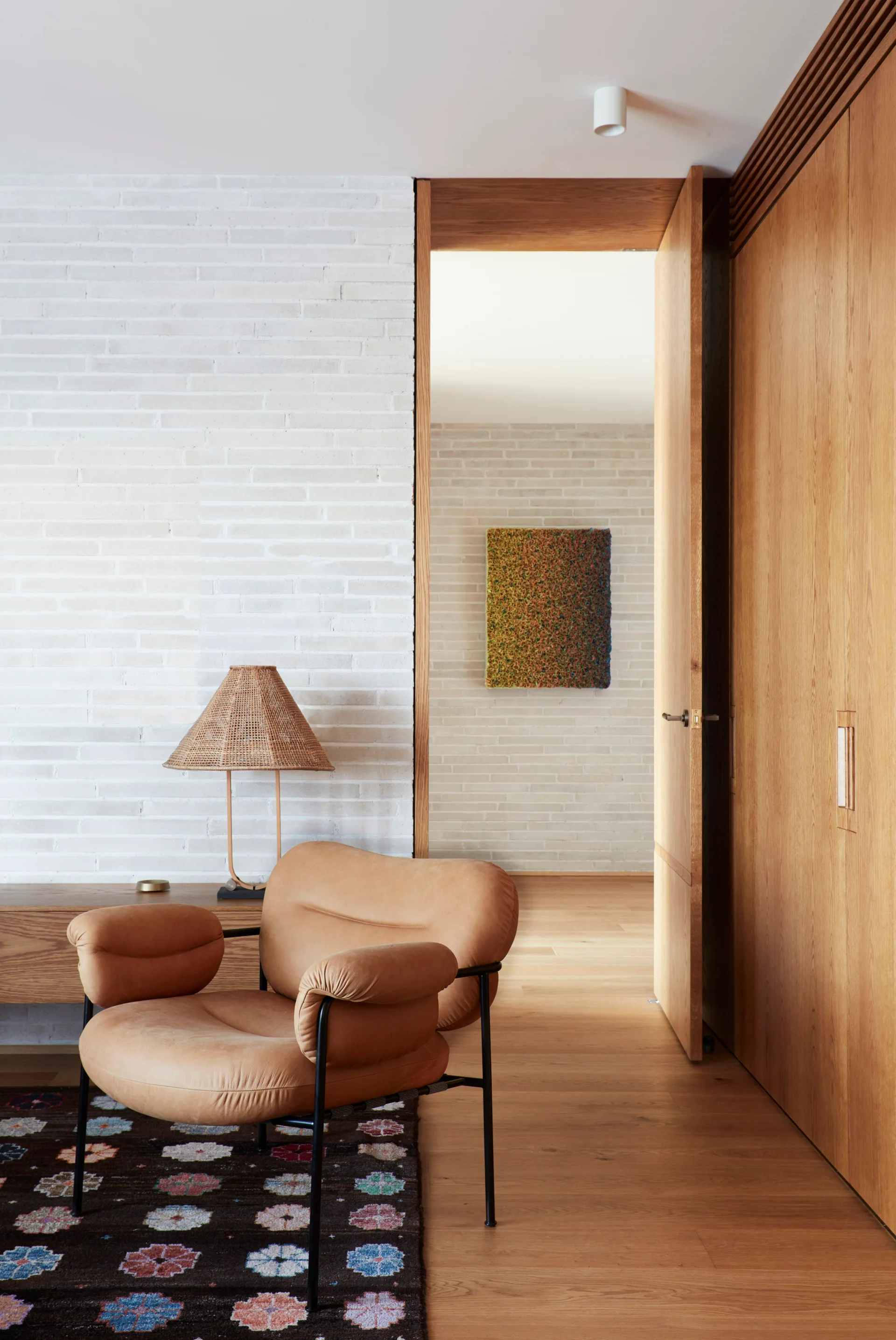 A tan leather armchair sits on a dark patterned rug in front of a white painted brick wall next to tall wooden closet doors.