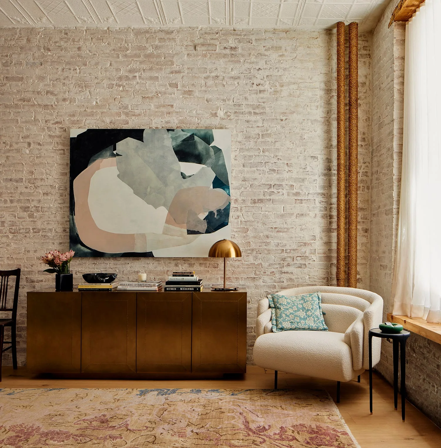 A brass console table and curved cream boucle armchair sit against a whitewashed brick wall beneath a large abstract painting.