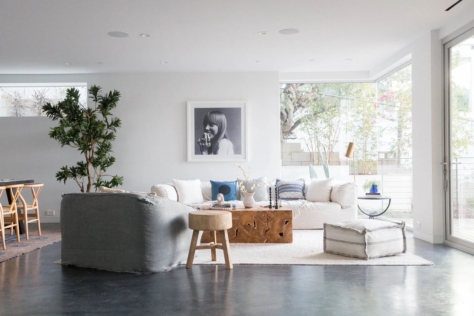 A bright living room features a white sofa, a carved wood block coffee table, a gray armchair, and large windows looking out to greenery.