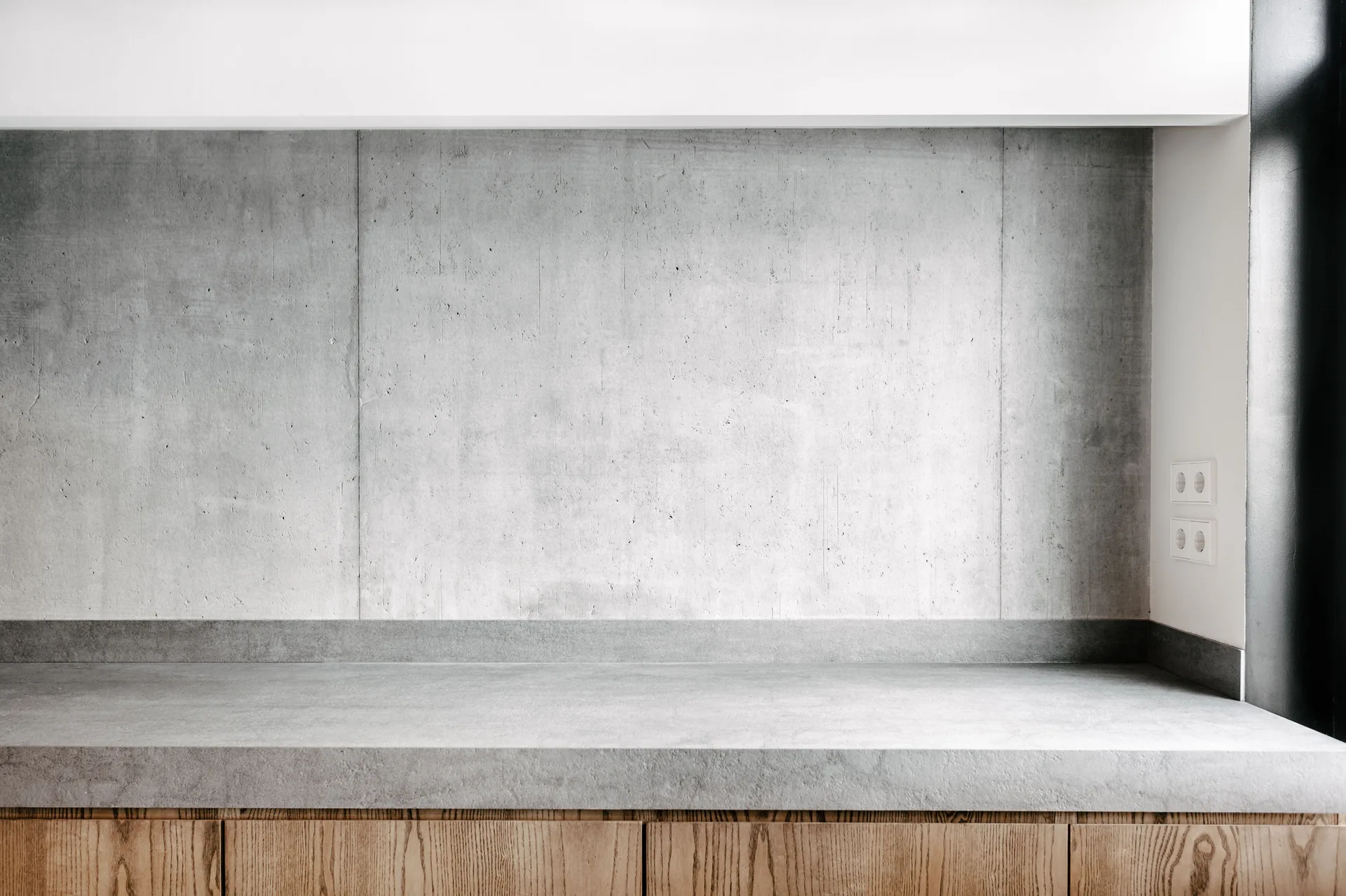 A minimalist kitchen view showing a gray concrete countertop and matching backsplash above wooden cabinetry with natural wood grain.