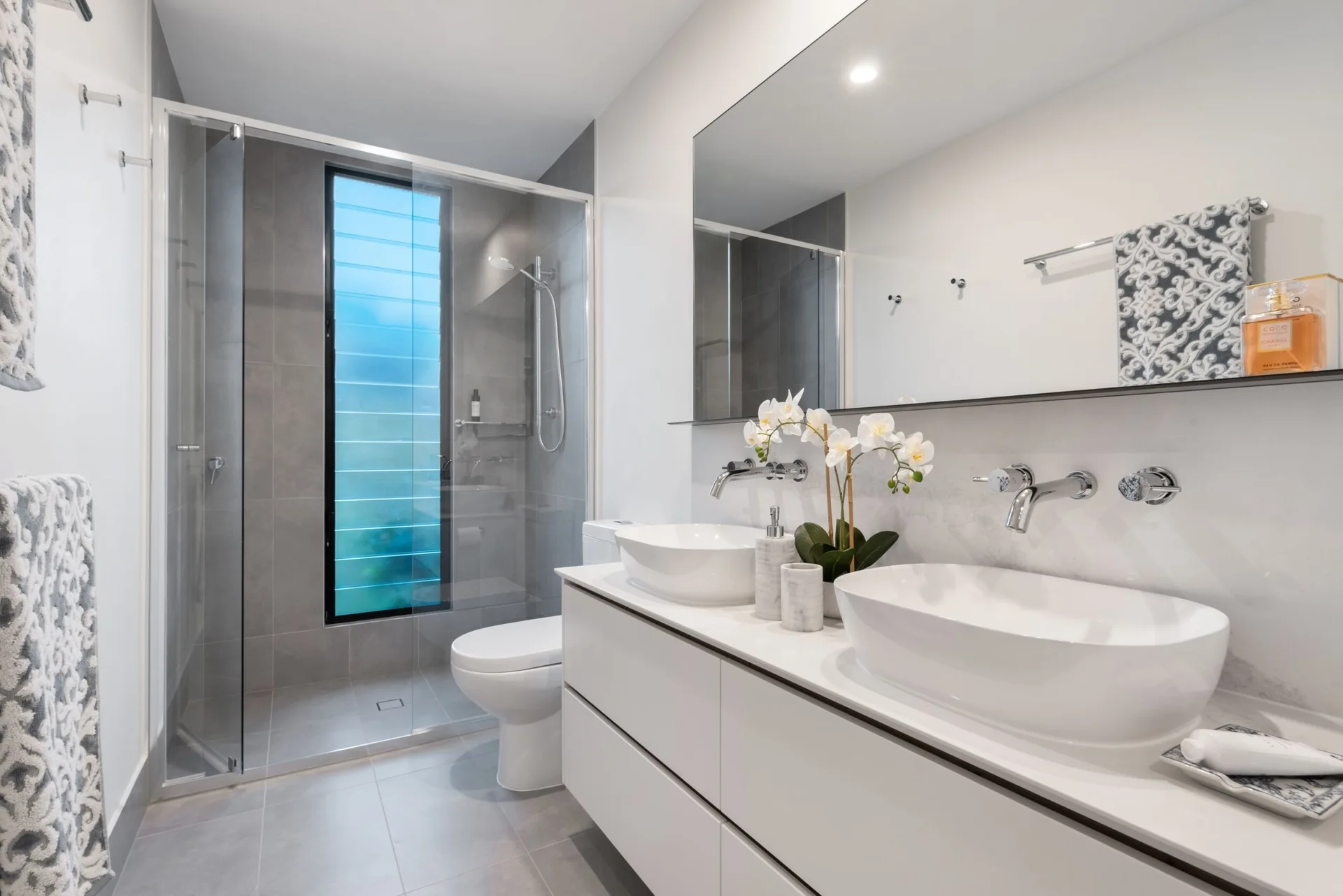 A modern bathroom featuring a white double vanity with vessel sinks, wall-mounted faucets, a large mirror, and a glass-enclosed walk-in shower.