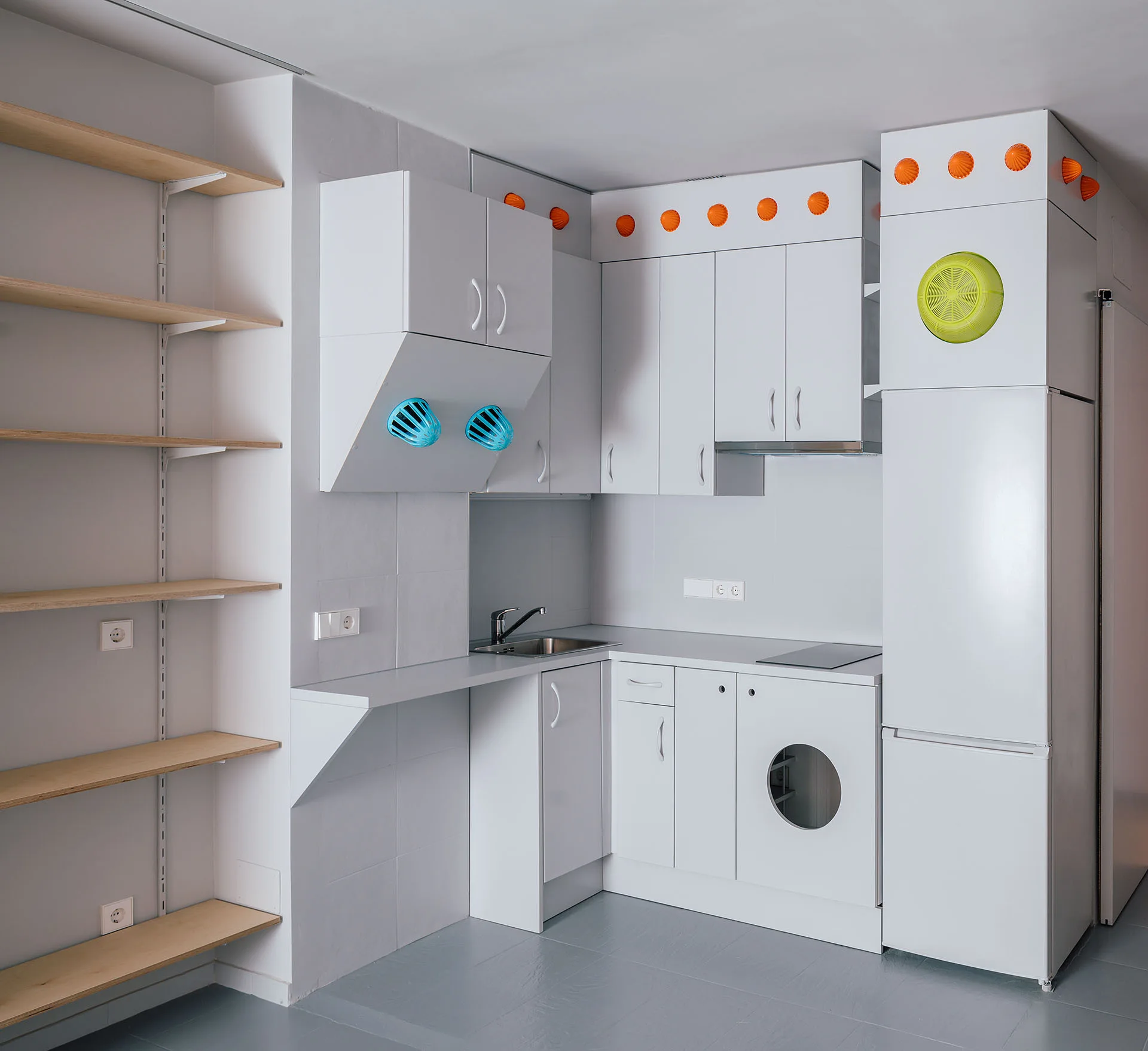 A compact white kitchen features matte cabinetry, an integrated washing machine, a refrigerator, and open wood-toned shelving on a grey floor.
