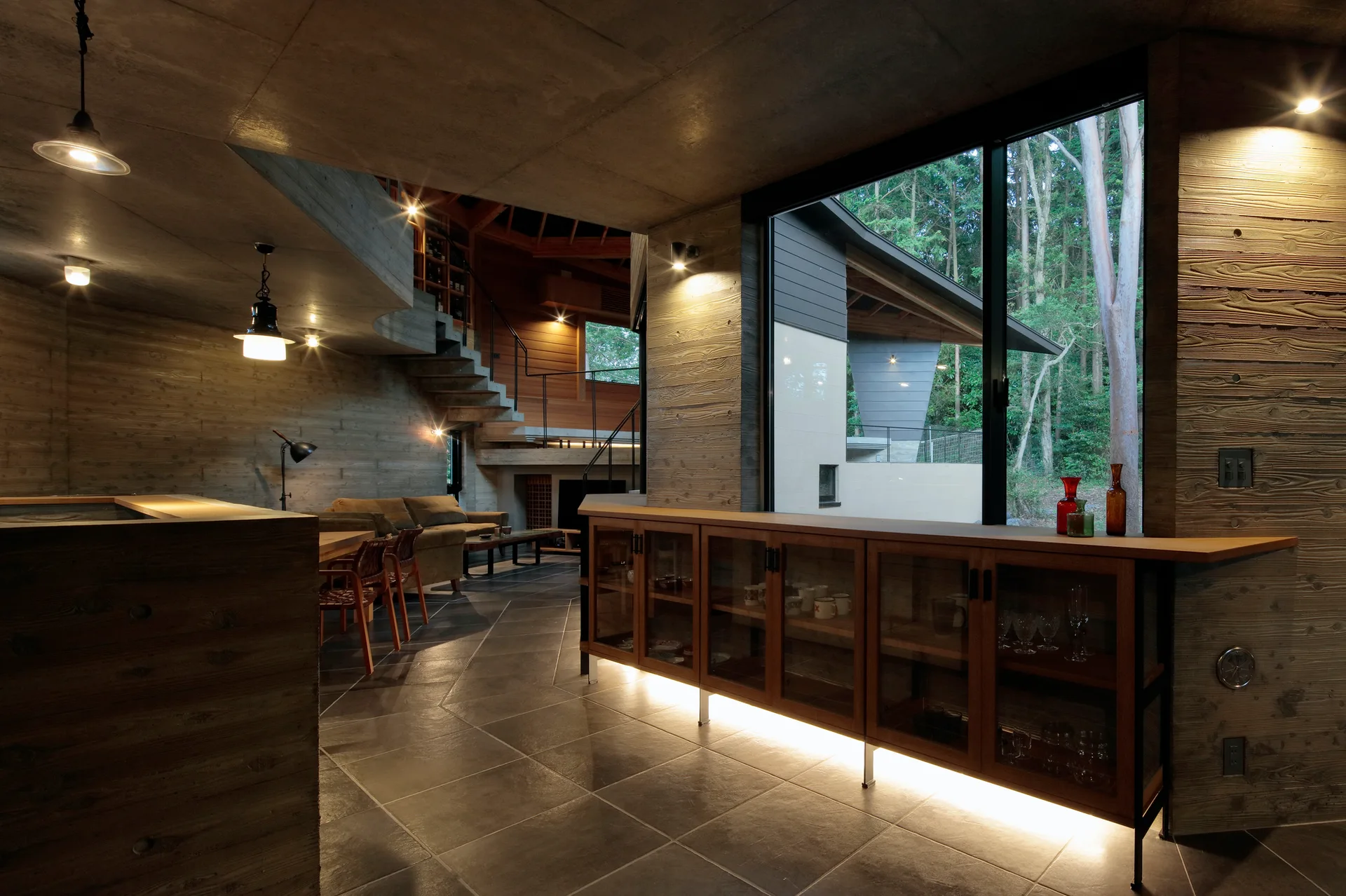 An open concrete interior features board-marked walls, a wood console cabinet with under-lighting, and a large window looking out onto a dense forest.