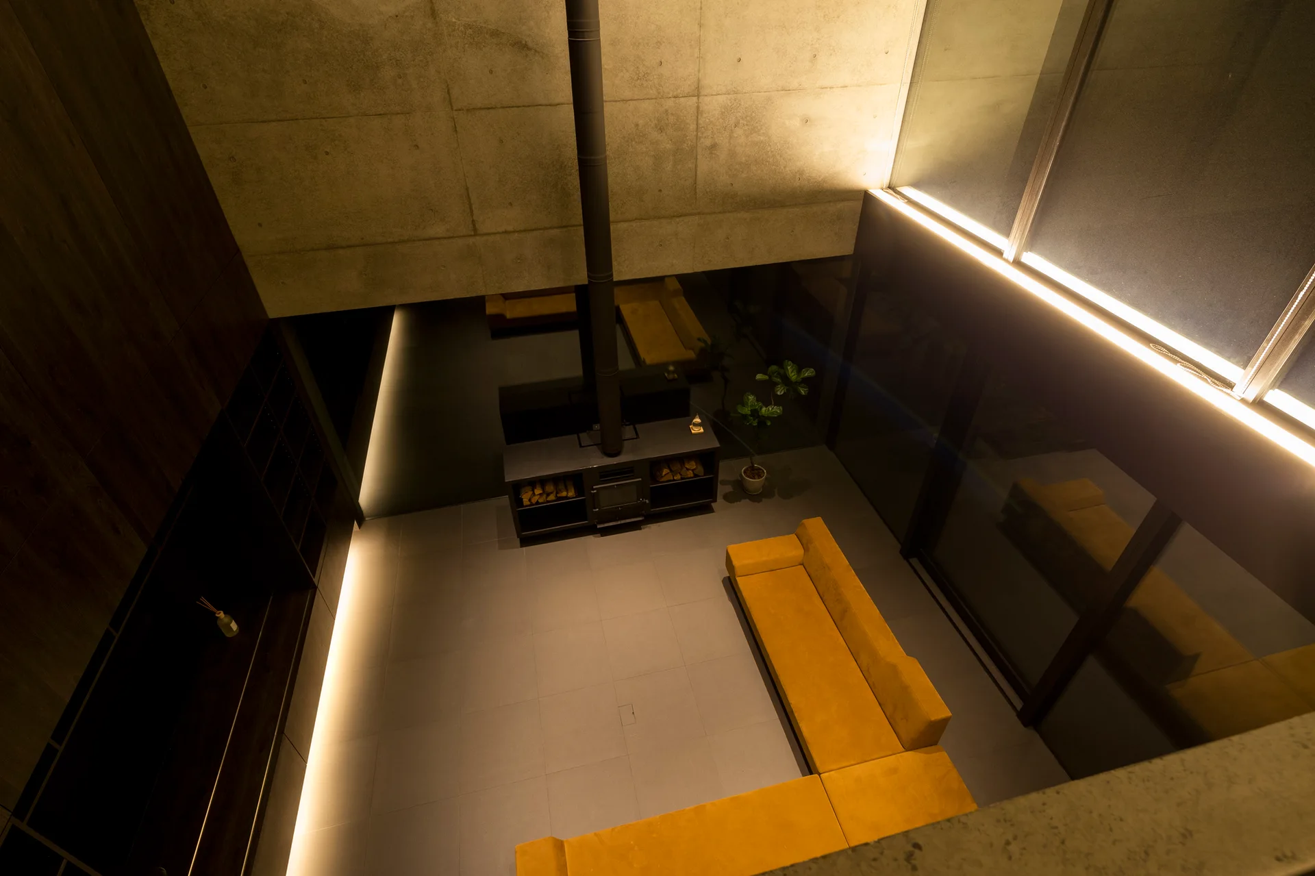 High-angle view of a minimalist living room featuring a yellow L-shaped sofa, a black wood-burning stove fireplace, and exposed concrete walls.