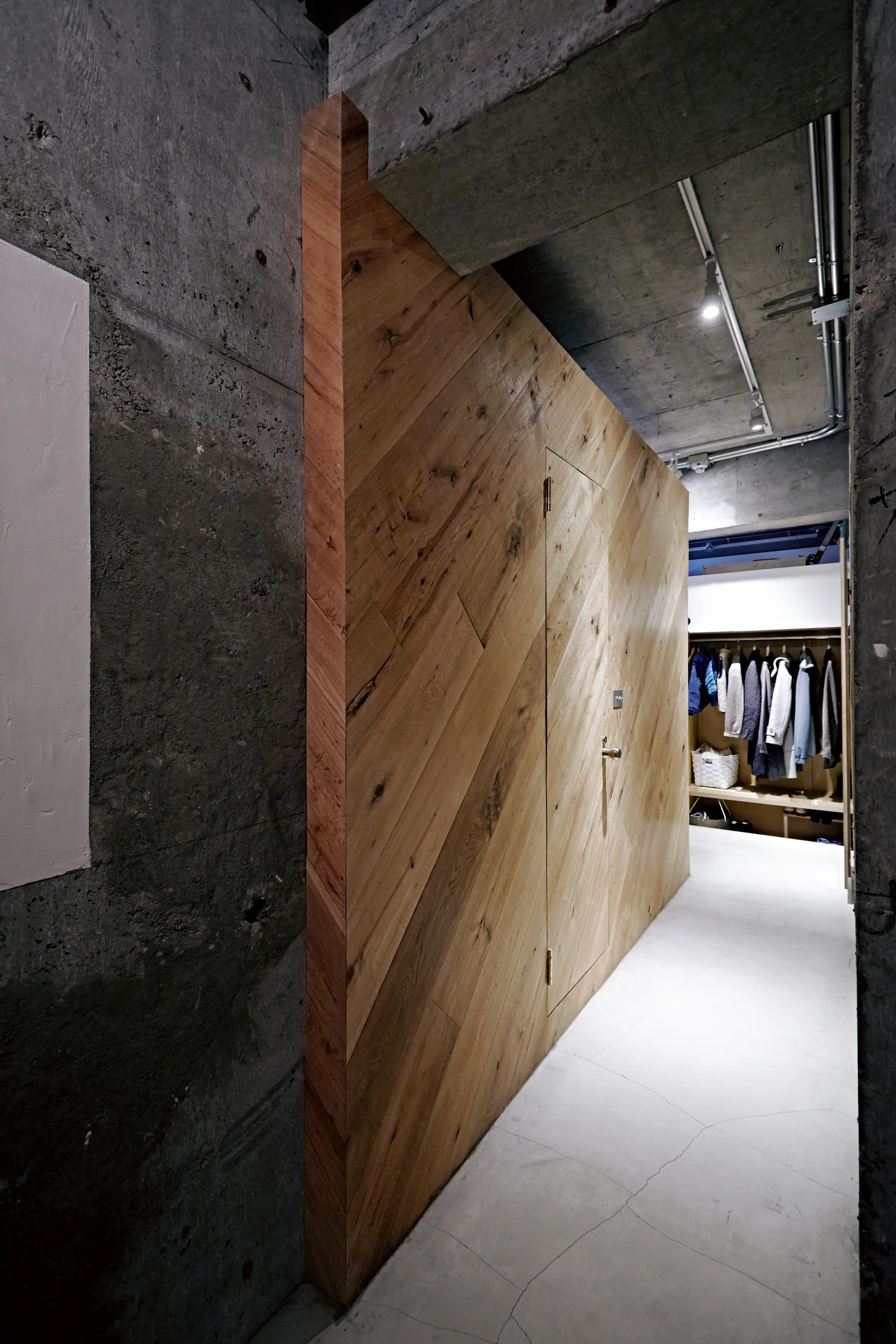 A hallway with exposed concrete walls and ceiling features a large partition wall clad in diagonal warm wood planks leading to a walk-in closet.