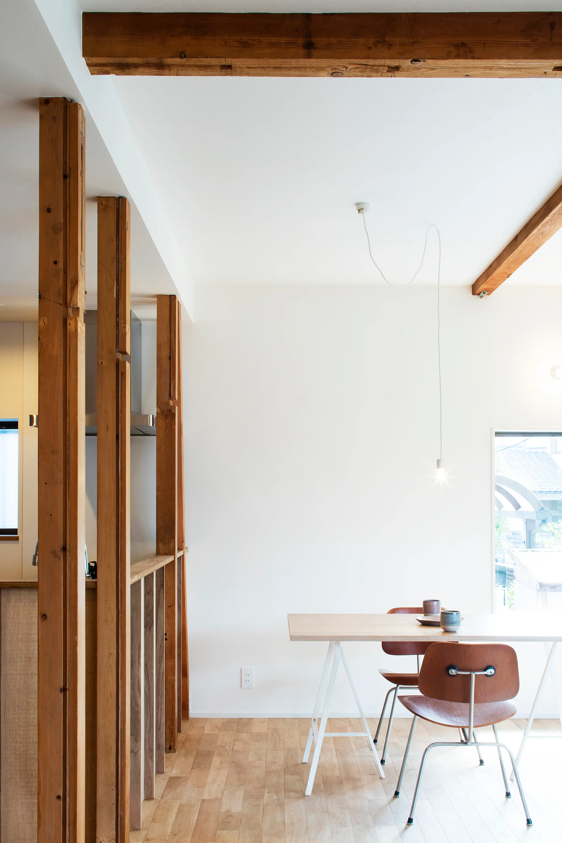 A minimalist dining area featuring a light wood trestle table, curved wooden chairs, and exposed raw timber pillars against white walls.