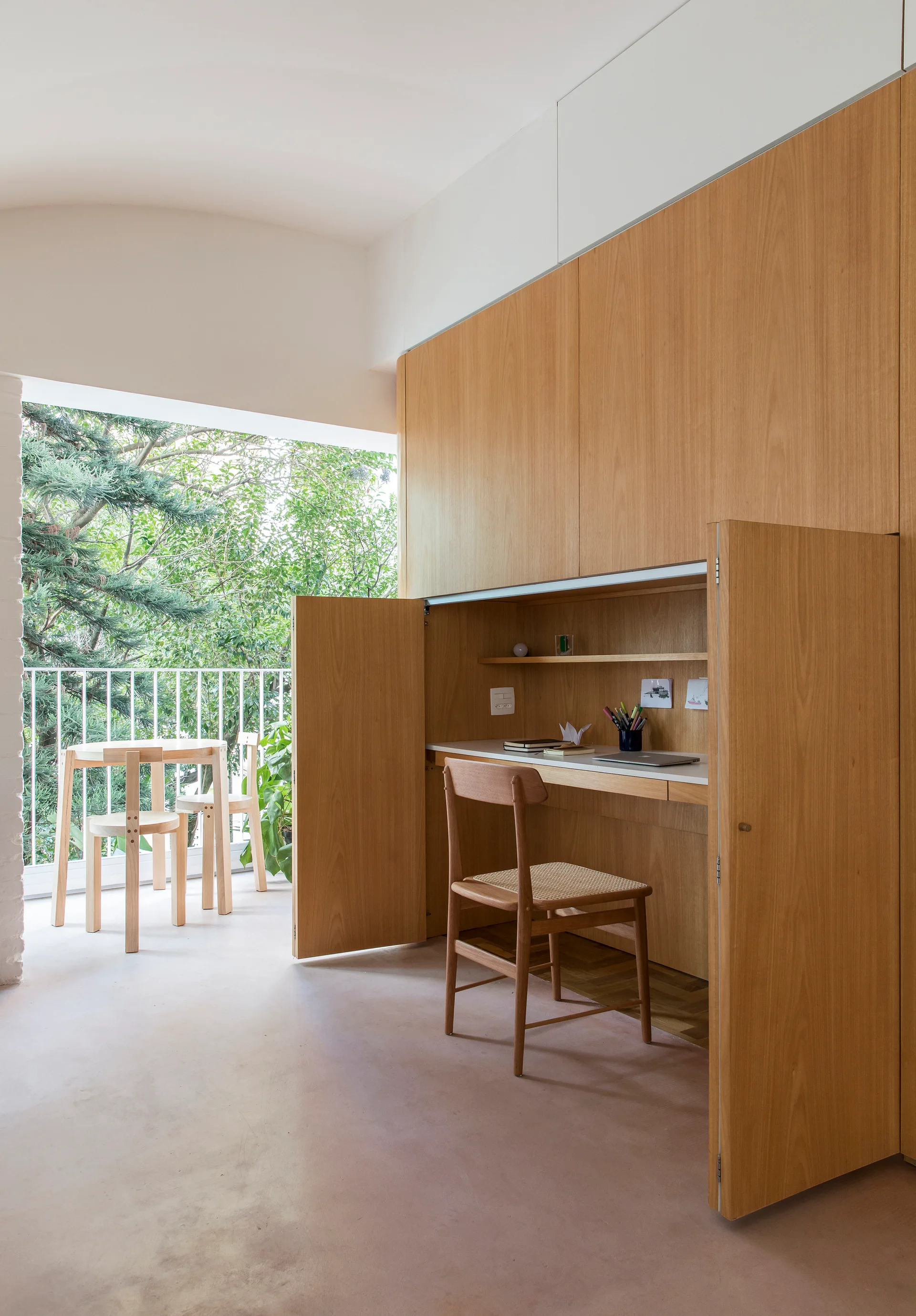 A home office workspace features an integrated wood desk built into large cabinetry, next to a balcony with a small table and chairs.