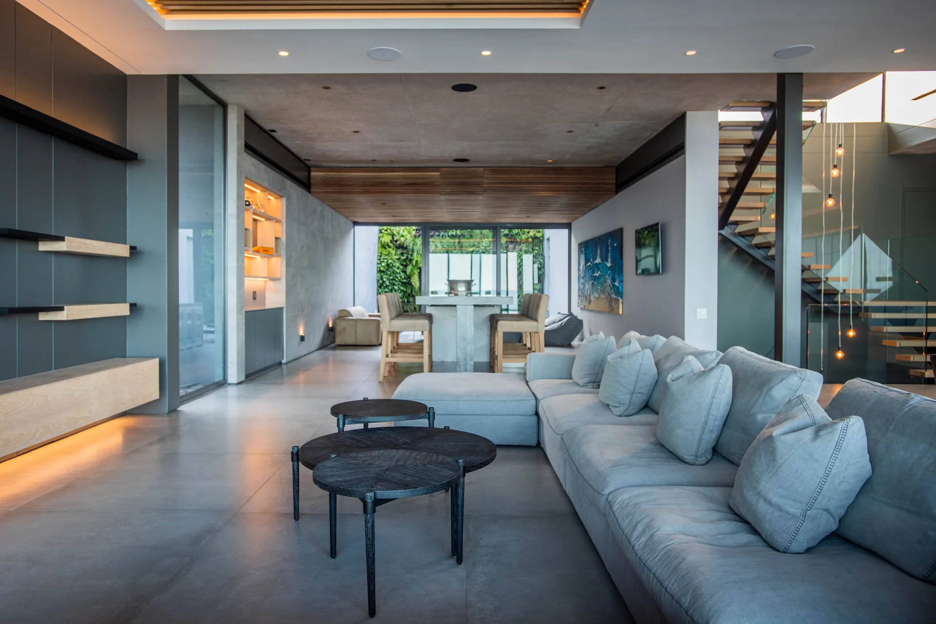 A spacious modern living room features a large light grey sectional sofa, nested dark wood coffee tables, and a floating staircase in the background.