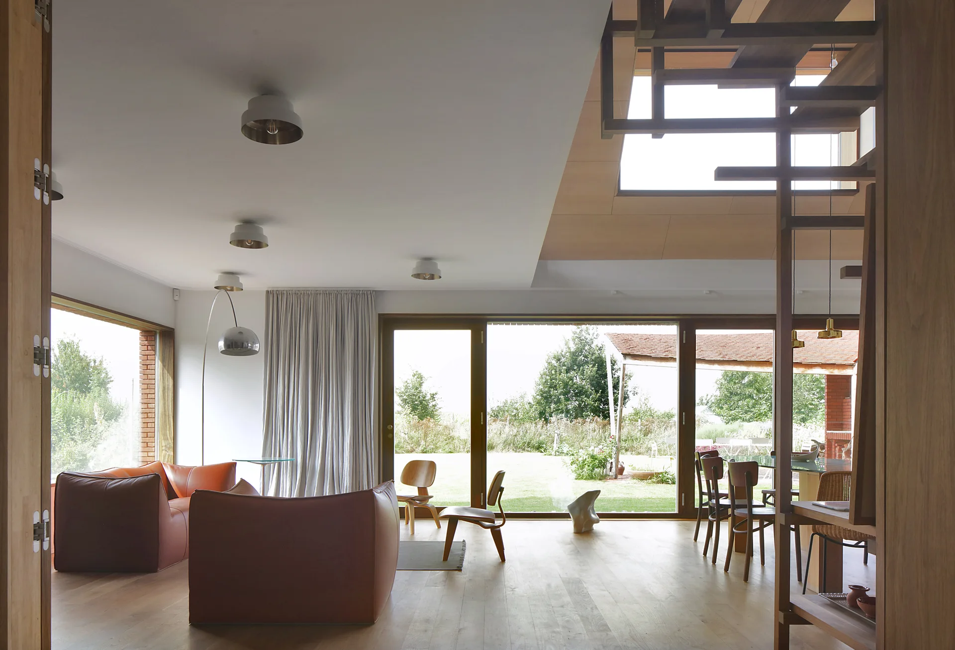An open-plan living room features low-profile brown leather sofas, wooden chairs, an open riser staircase, and large sliding glass doors opening to a yard.