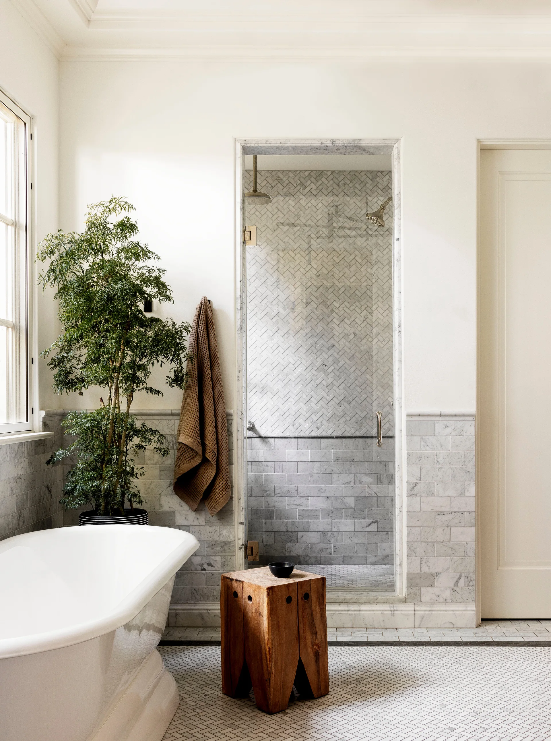 A bright bathroom with a white freestanding tub, a rustic wooden stool, a large green potted plant, and a tiled walk-in shower.